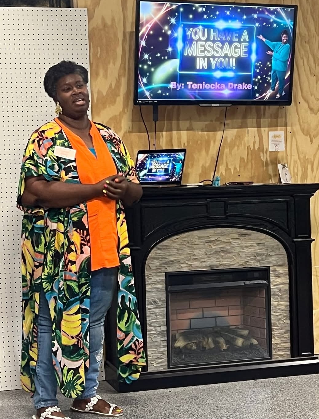 Person standing in front of a fireplace with a colorful robe, orange shirt, and jeans, smiling at the camera.