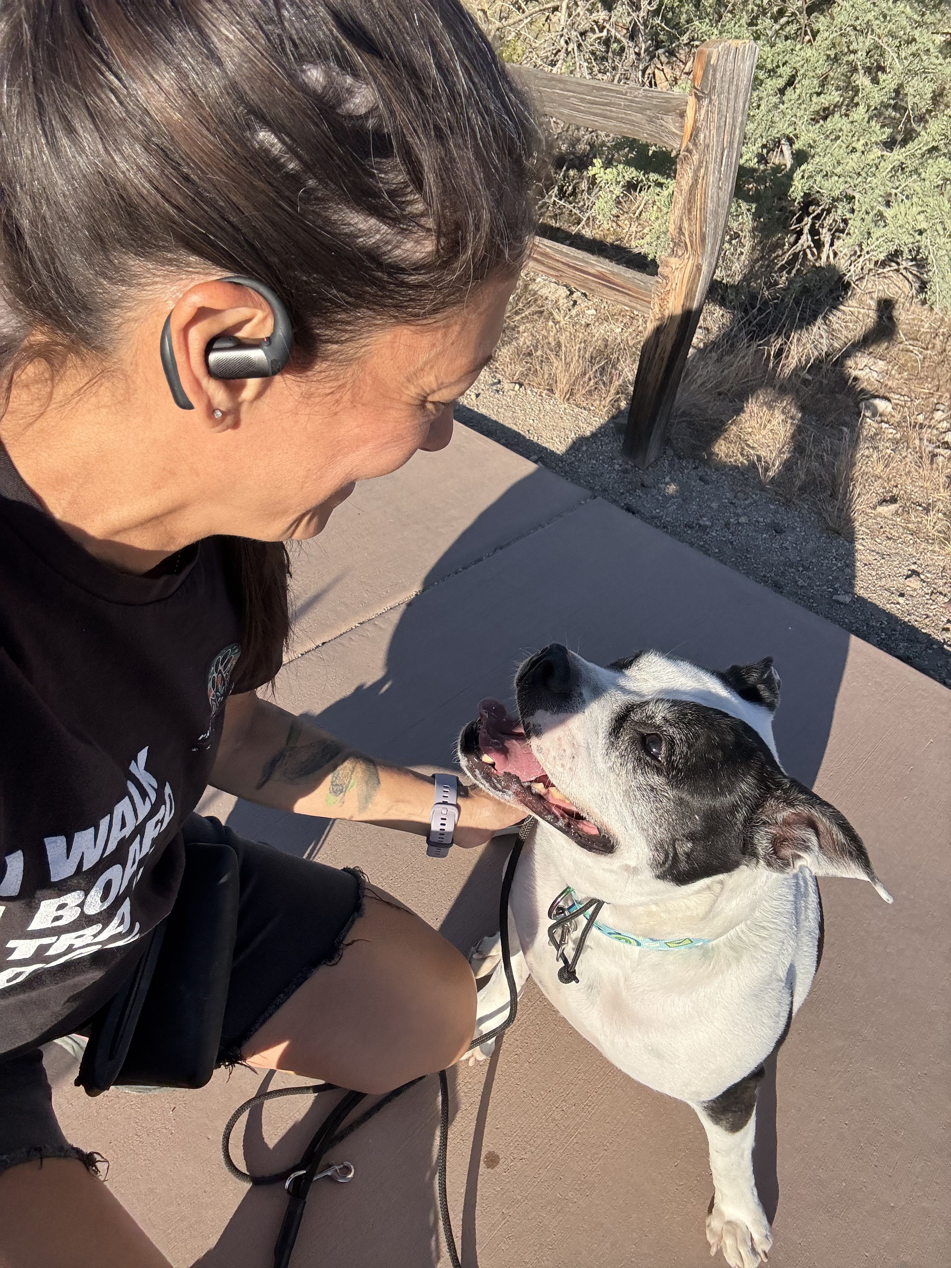 Woman with dark hair and earbud smiling at a black and white dog outdoors on a leash.