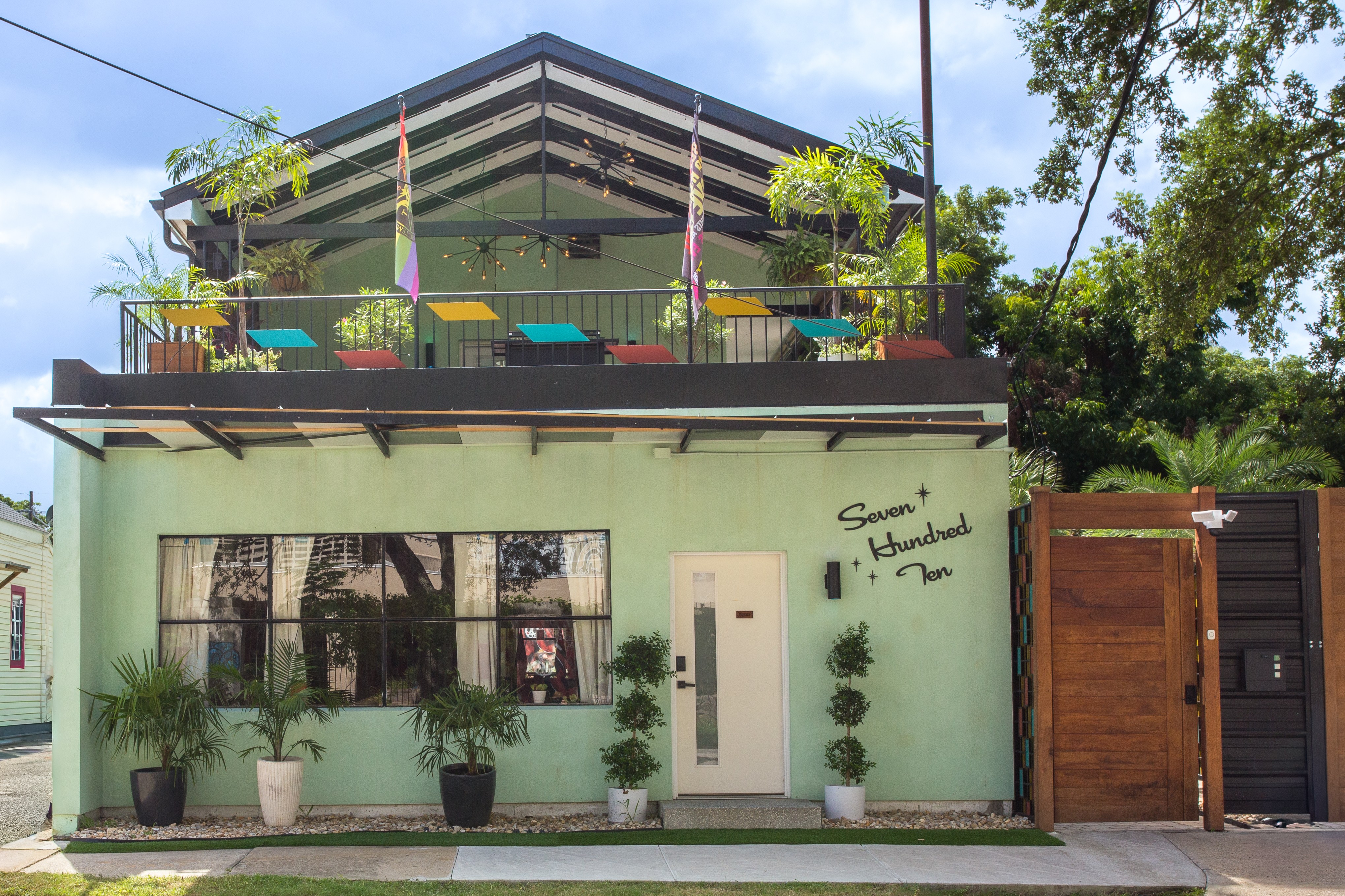 Two-story house with a green exterior, balcony with colorful chairs, potted plants outside, and a wooden gate on the right.