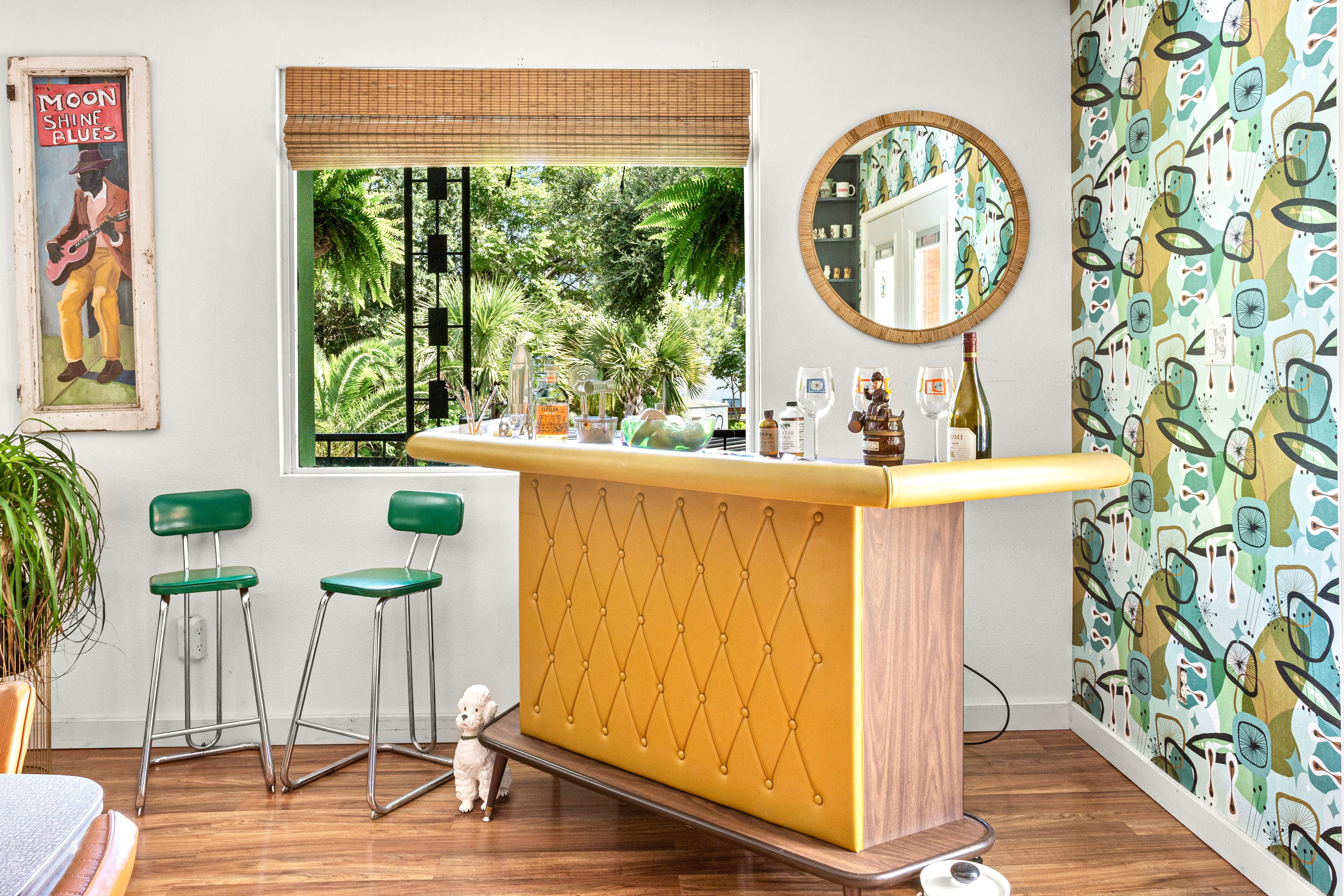 Interior with window, two green chairs, yellow bar, round mirror, patterned wallpaper, and plants.