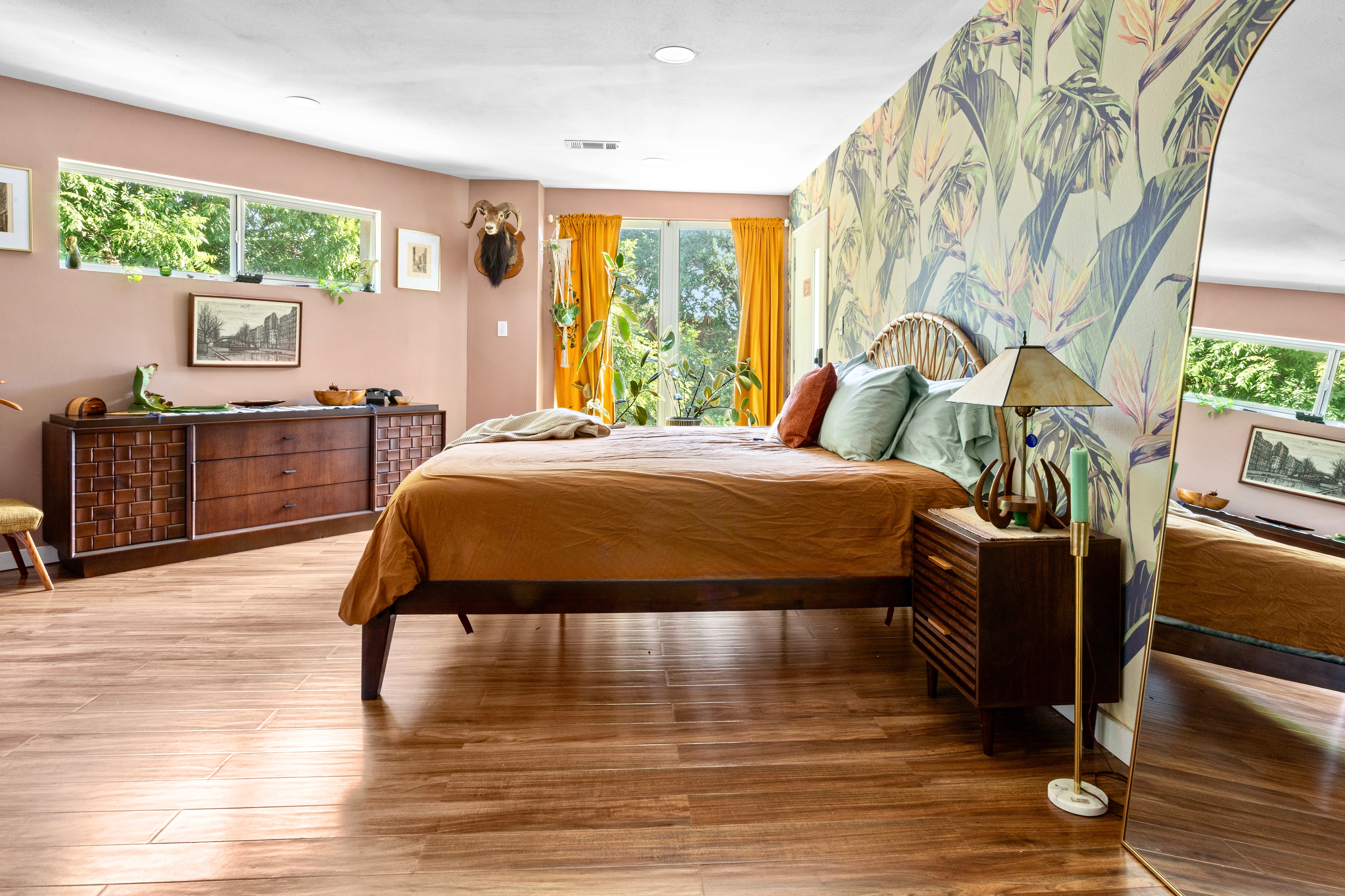 Bedroom with large bed, wooden furniture, colorful curtains, and a floral wall design, with windows showing greenery outside.