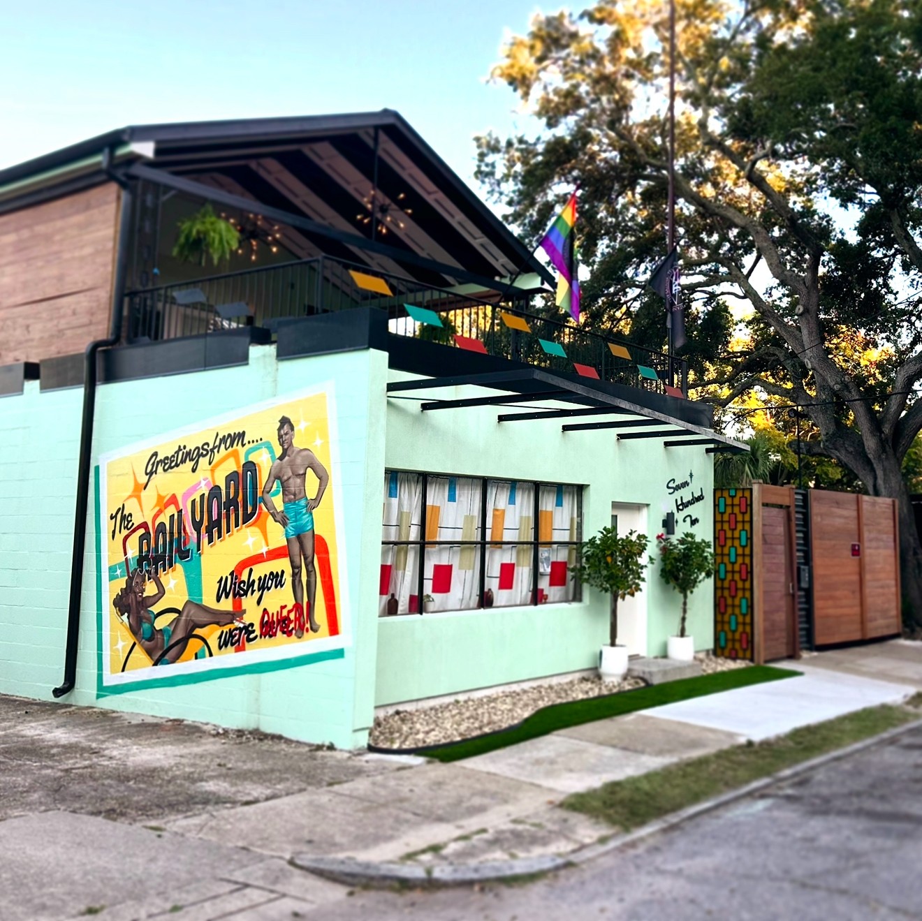 Building with a colorful mural, windows with red accents, potted plants, and a rainbow flag, surrounded by trees and sidewalk.