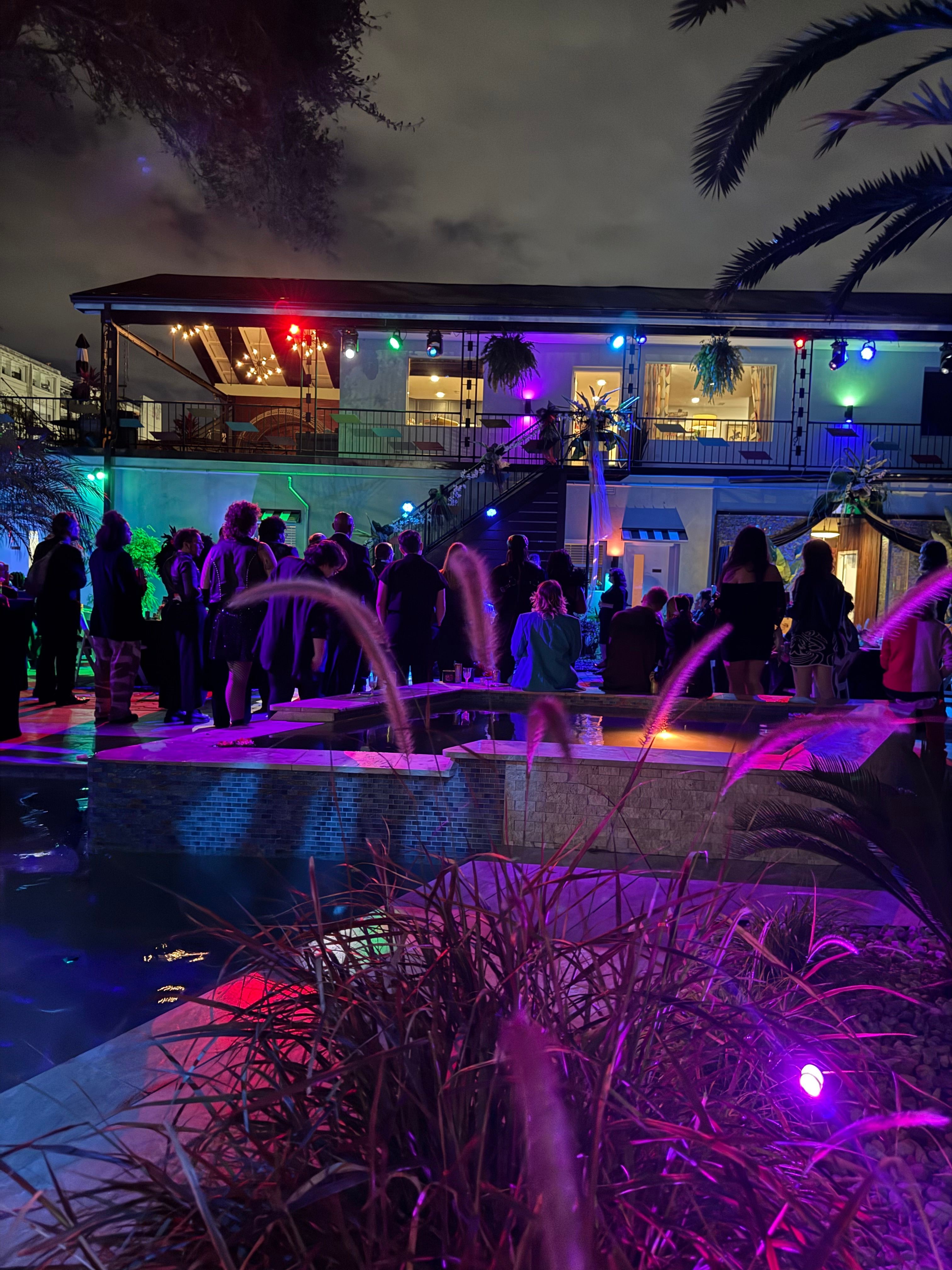 Night scene at a poolside party with colorful lights, people gathered, and a building in the background.