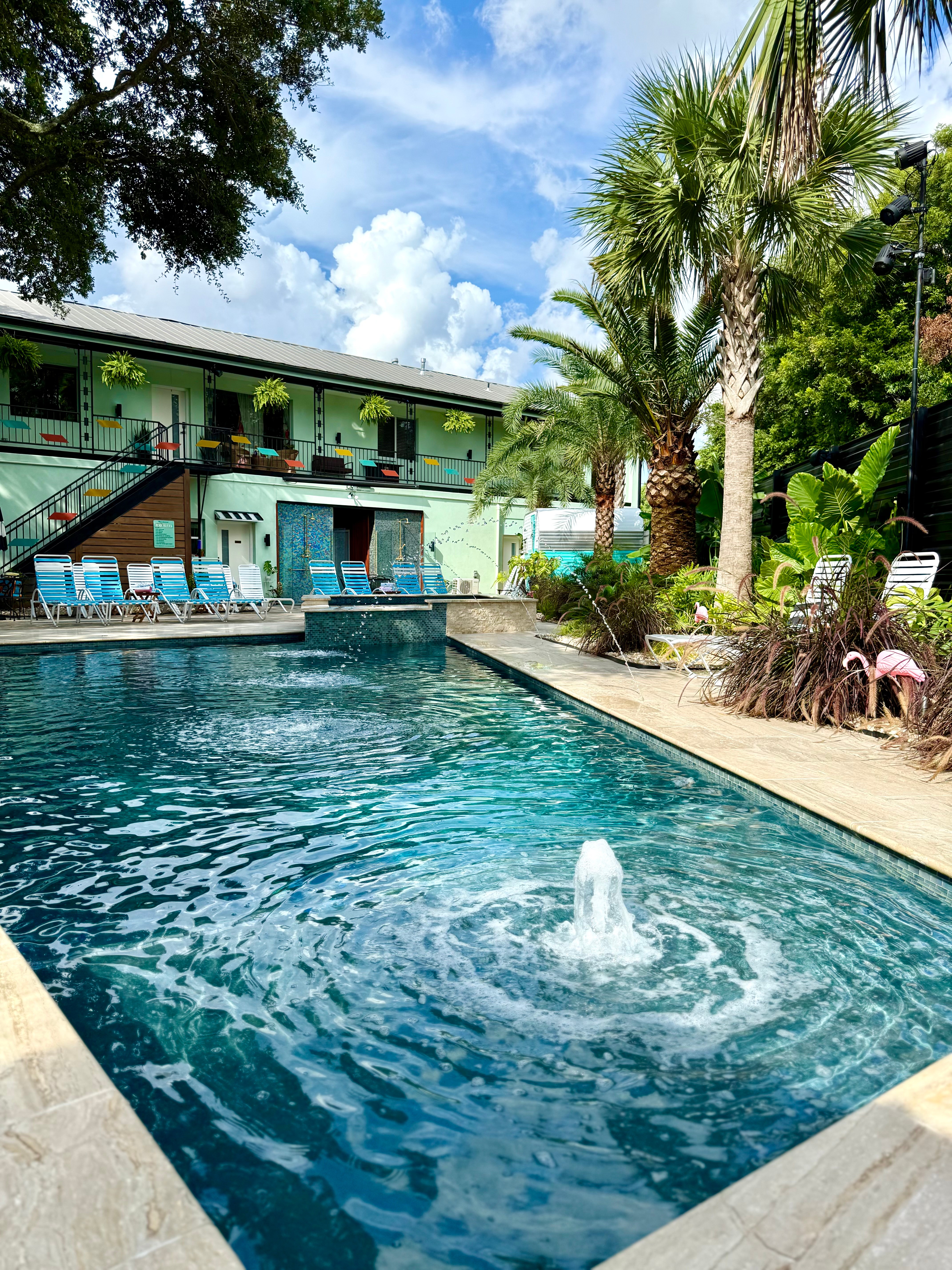 Swimming pool with water fountain, surrounded by lounge chairs, plants, and a two-story building with balconies under a partly cloudy sky.