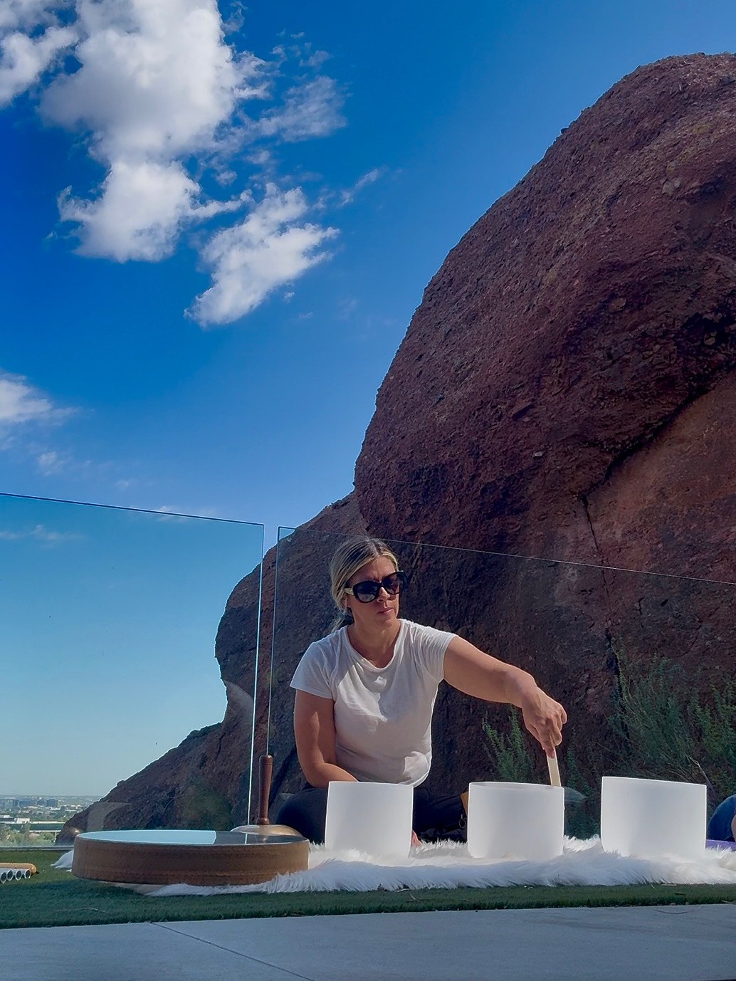 Woman wearing sunglasses reaching for white objects on table outdoors with large rock and blue sky in background.