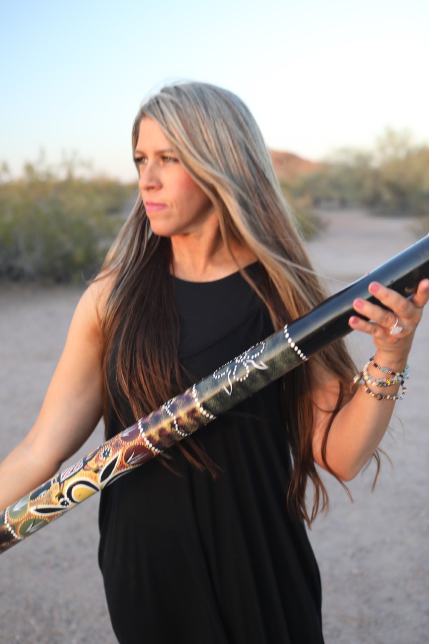Woman with long hair holding a didgeridoo outdoors, wearing a black dress and bracelets, with a natural background.