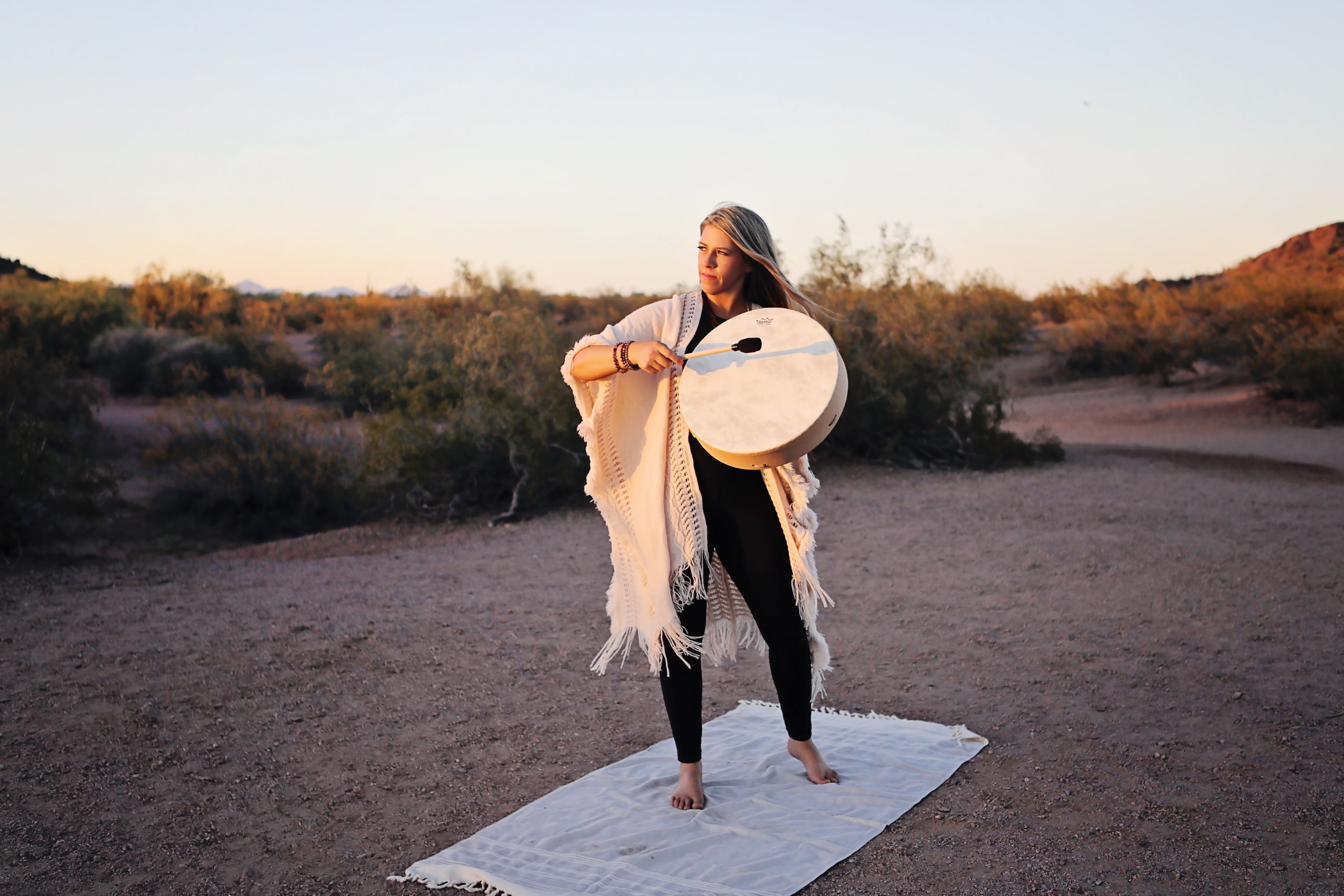 Person standing on a mat outdoors holding a large circular object, with desert landscape and bushes in background.