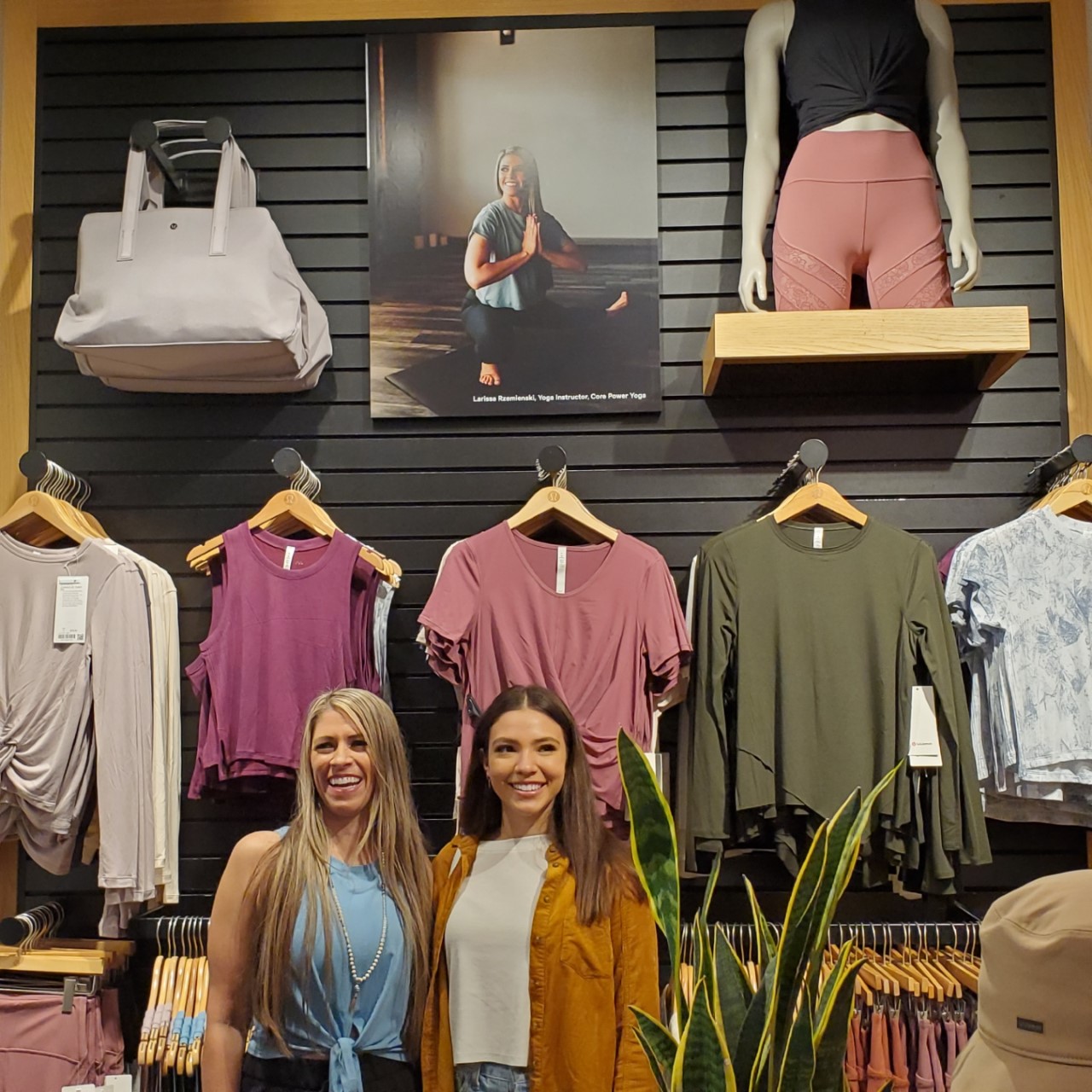 Two women smiling in a clothing store with various garments and mannequins displayed behind them.