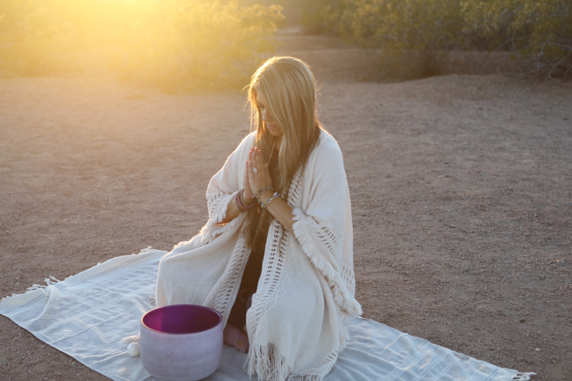 Person sitting on a blanket outdoors with hands in prayer position, sunlight shining from behind.