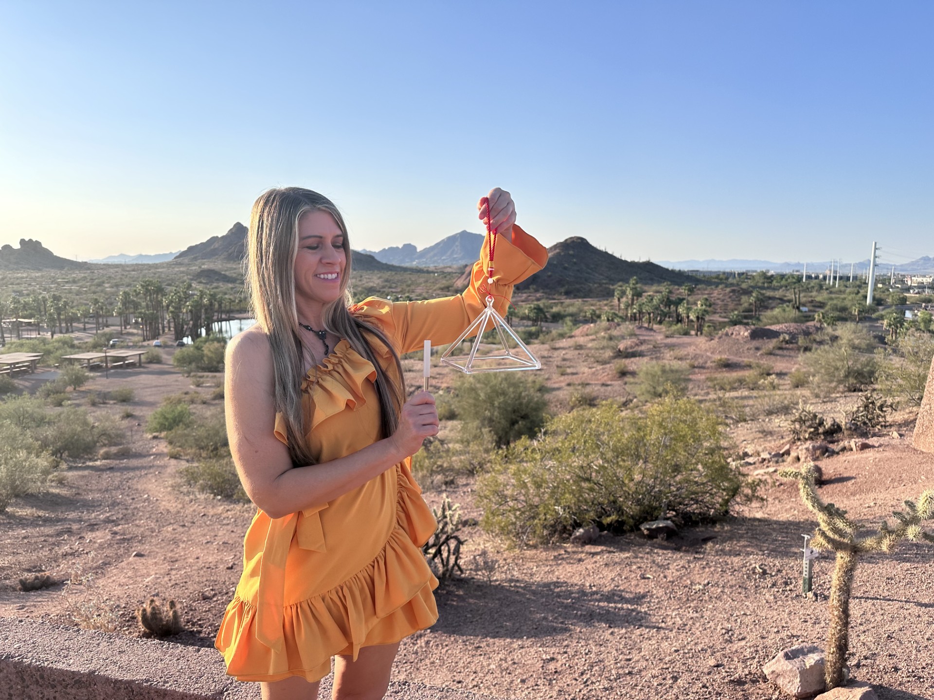 Woman in orange dress holding a doll in a desert landscape with mountains and sparse vegetation.