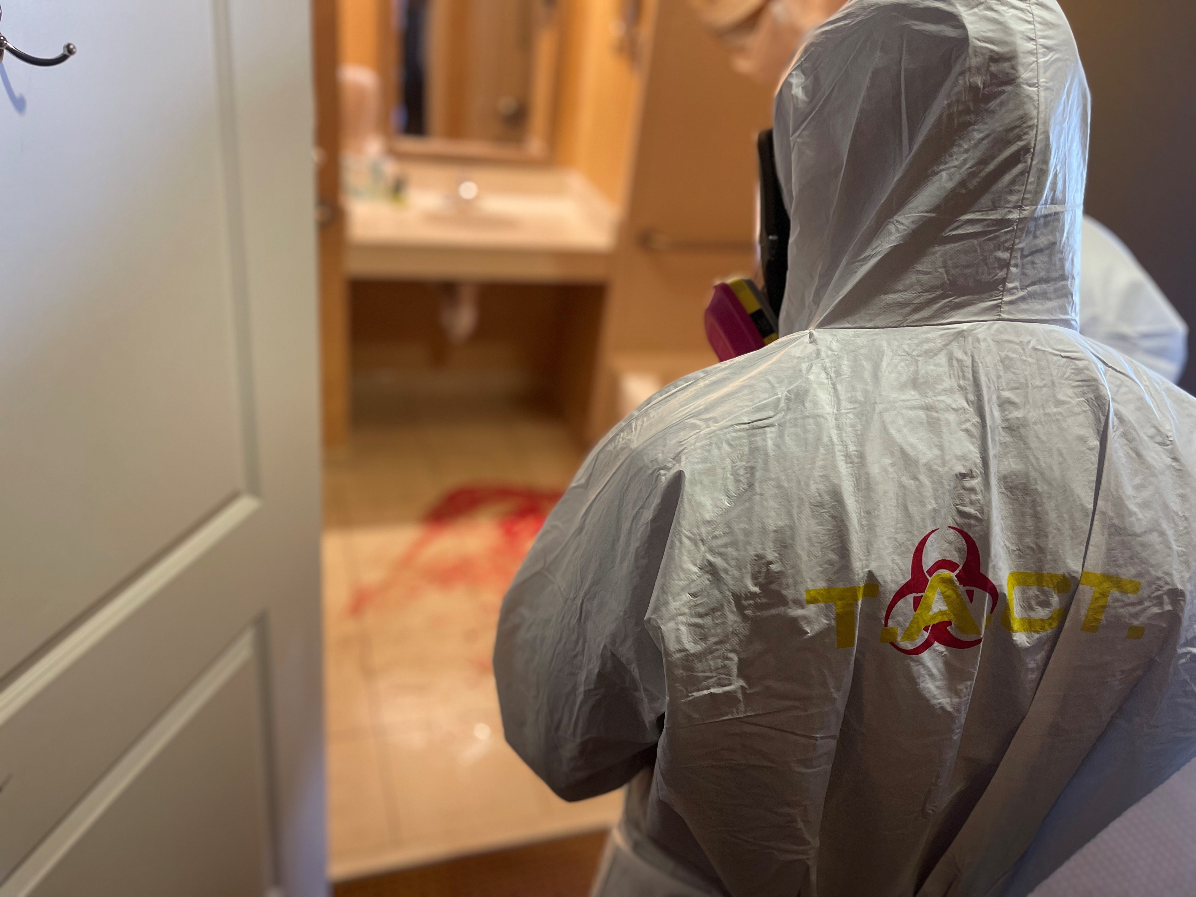 Person wearing a protective suit with a biohazard symbol on the back, facing away, in a room with a mirror and a red rug.
