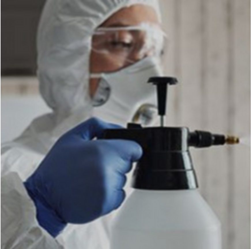 Person in protective gear holding a spray device, wearing gloves, goggles, and a mask.