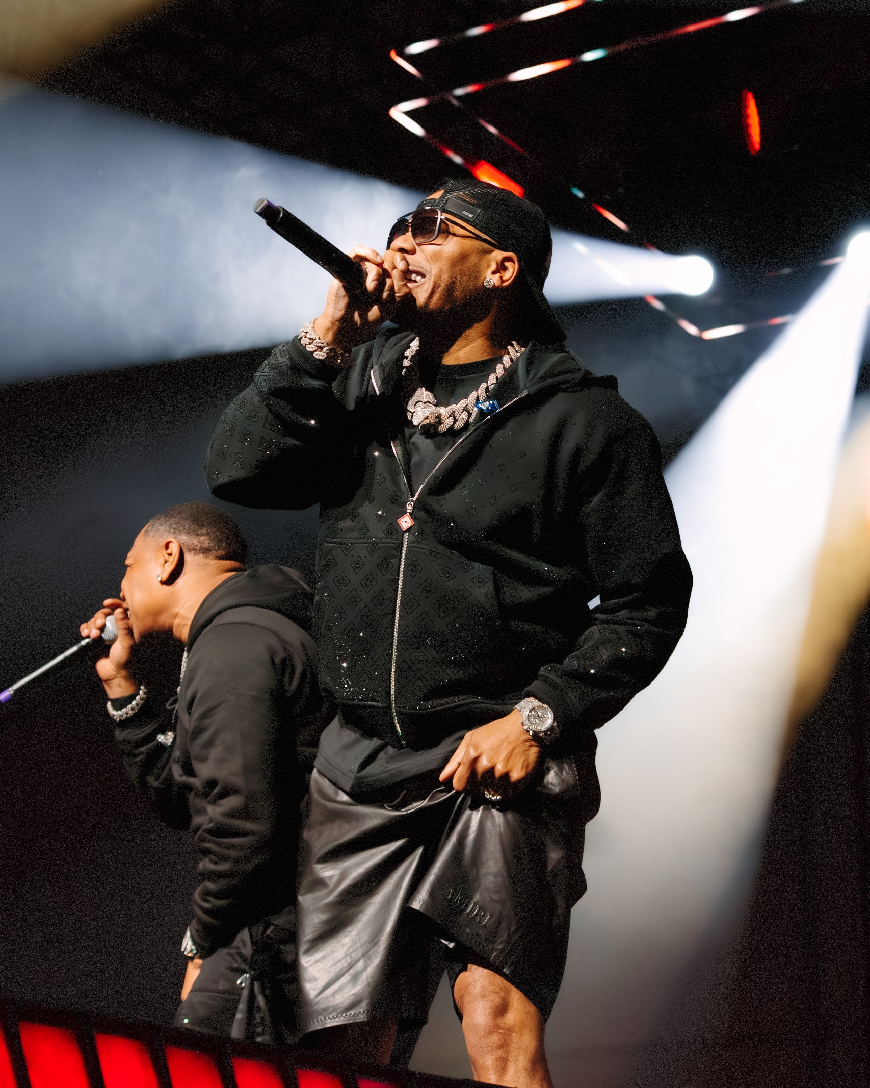 Performer with sunglasses singing into microphone on stage with bright lights and smoke effects.