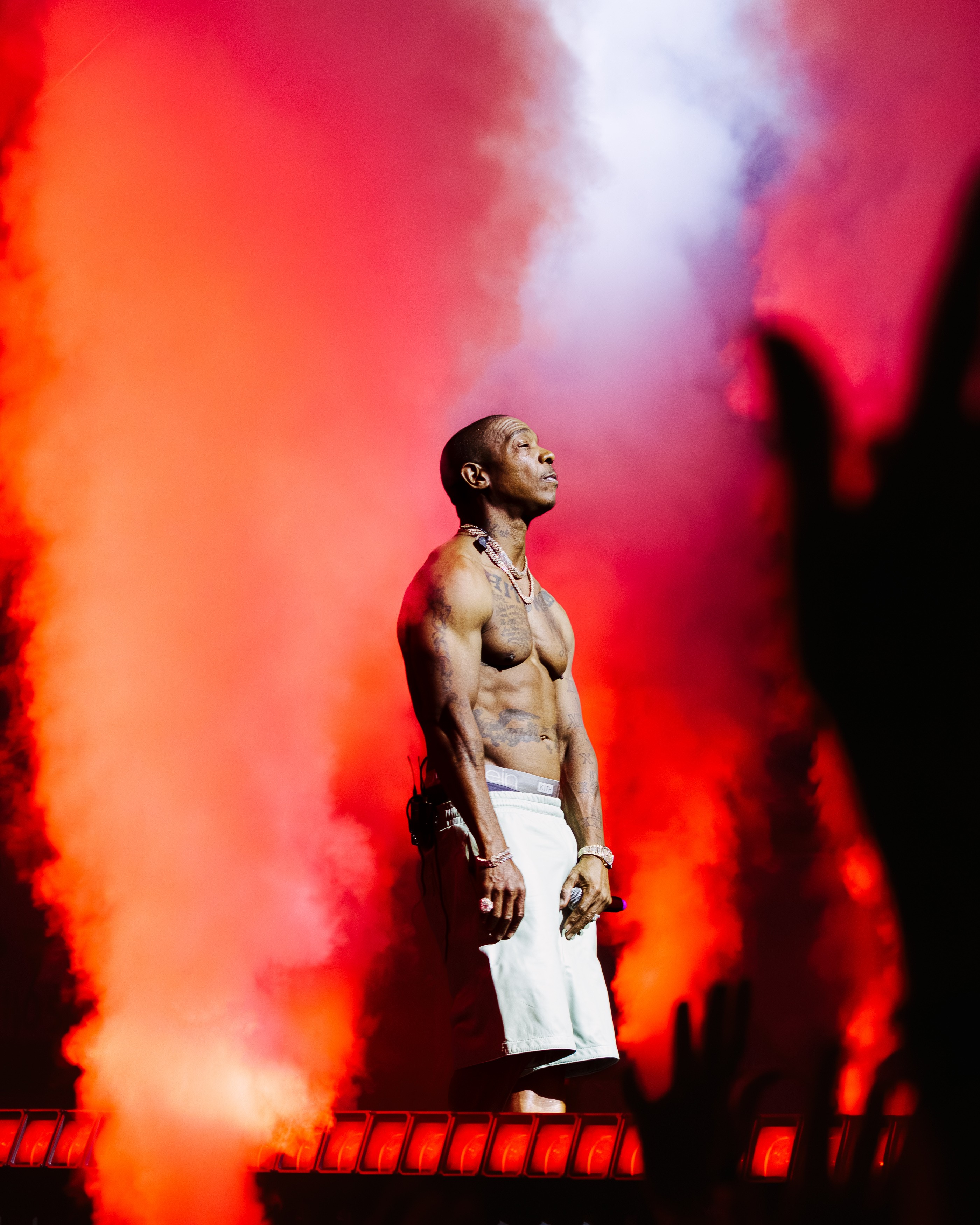 Male performer on stage with colorful smoke and lighting, shirtless, wearing white pants, holding a microphone, looking upward.