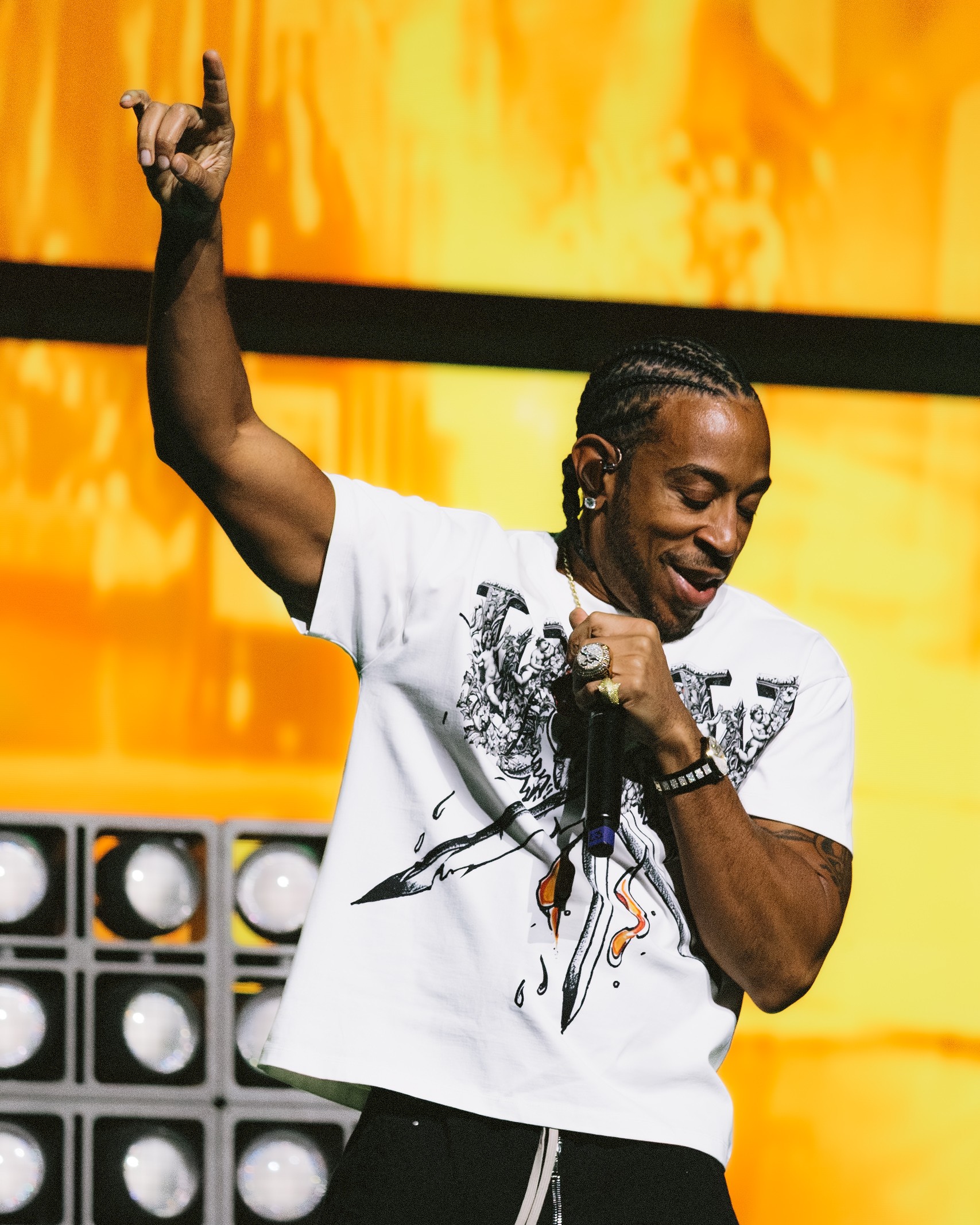 Person with braided hair smiling, holding a microphone, raising one arm, in front of a yellow background.