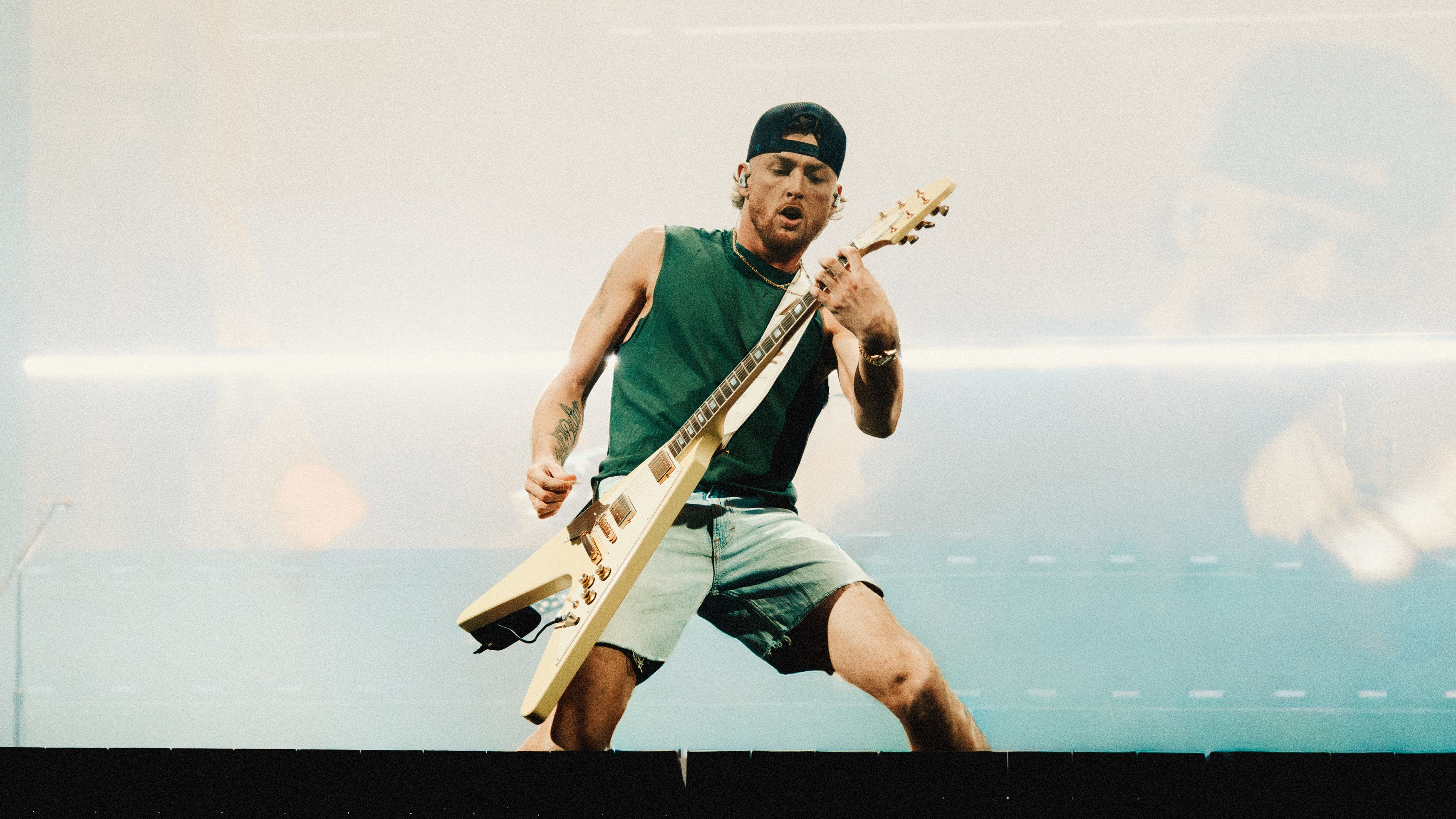 Person playing an electric guitar on stage, wearing a sleeveless shirt, shorts, and a cap.