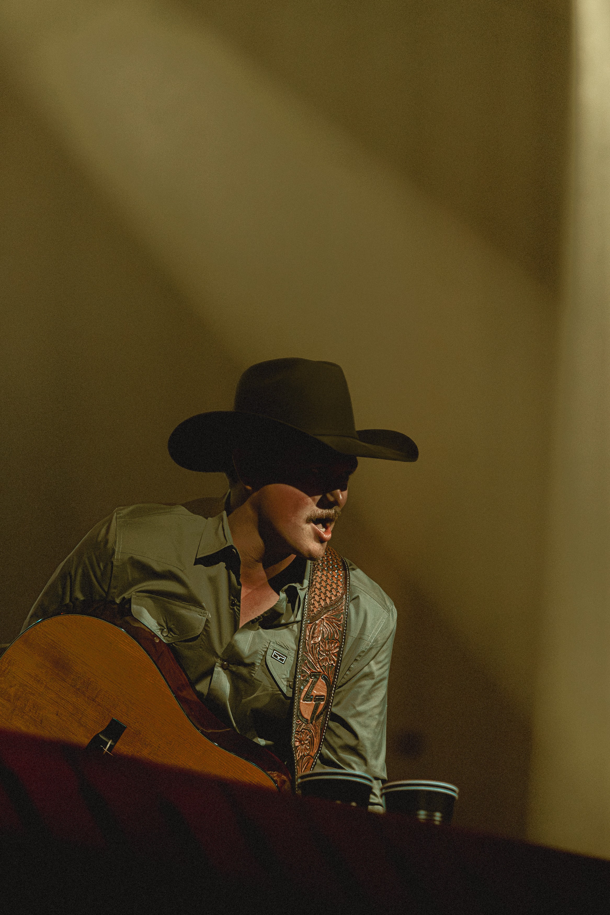 Person wearing a wide-brimmed hat and sunglasses, playing guitar on stage with a spotlight shining down.