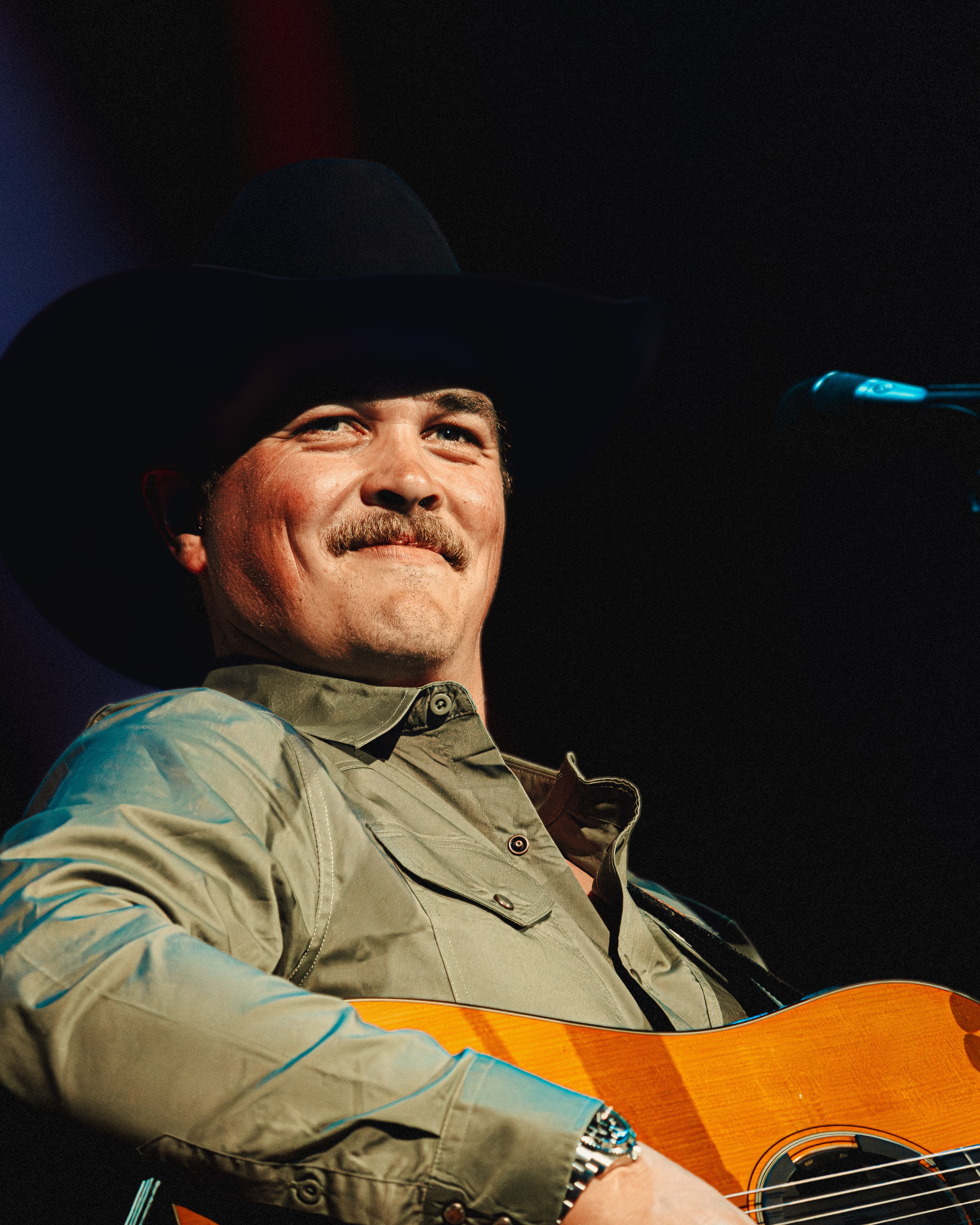 Man wearing a hat and shirt, smiling, holding a guitar, on stage with a dark background.