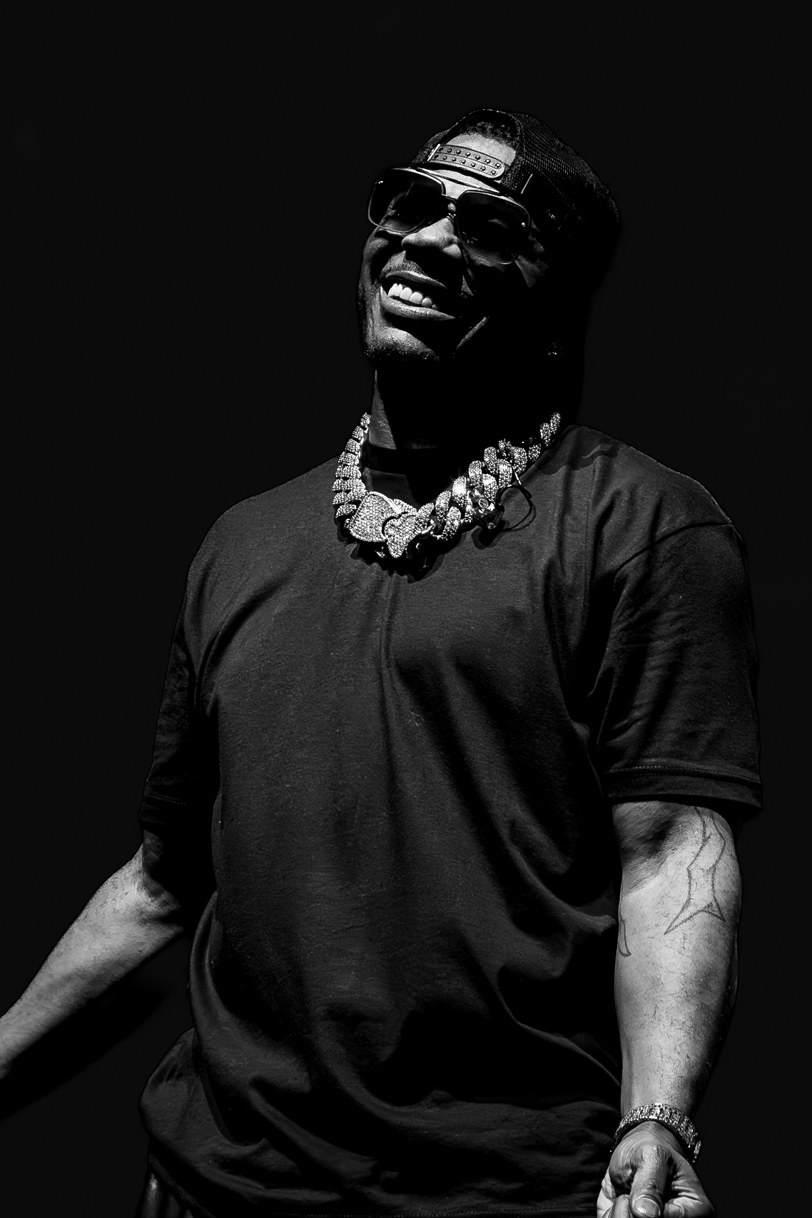 Person smiling, wearing sunglasses, a cap, a necklace, and a dark t-shirt, against a dark background.