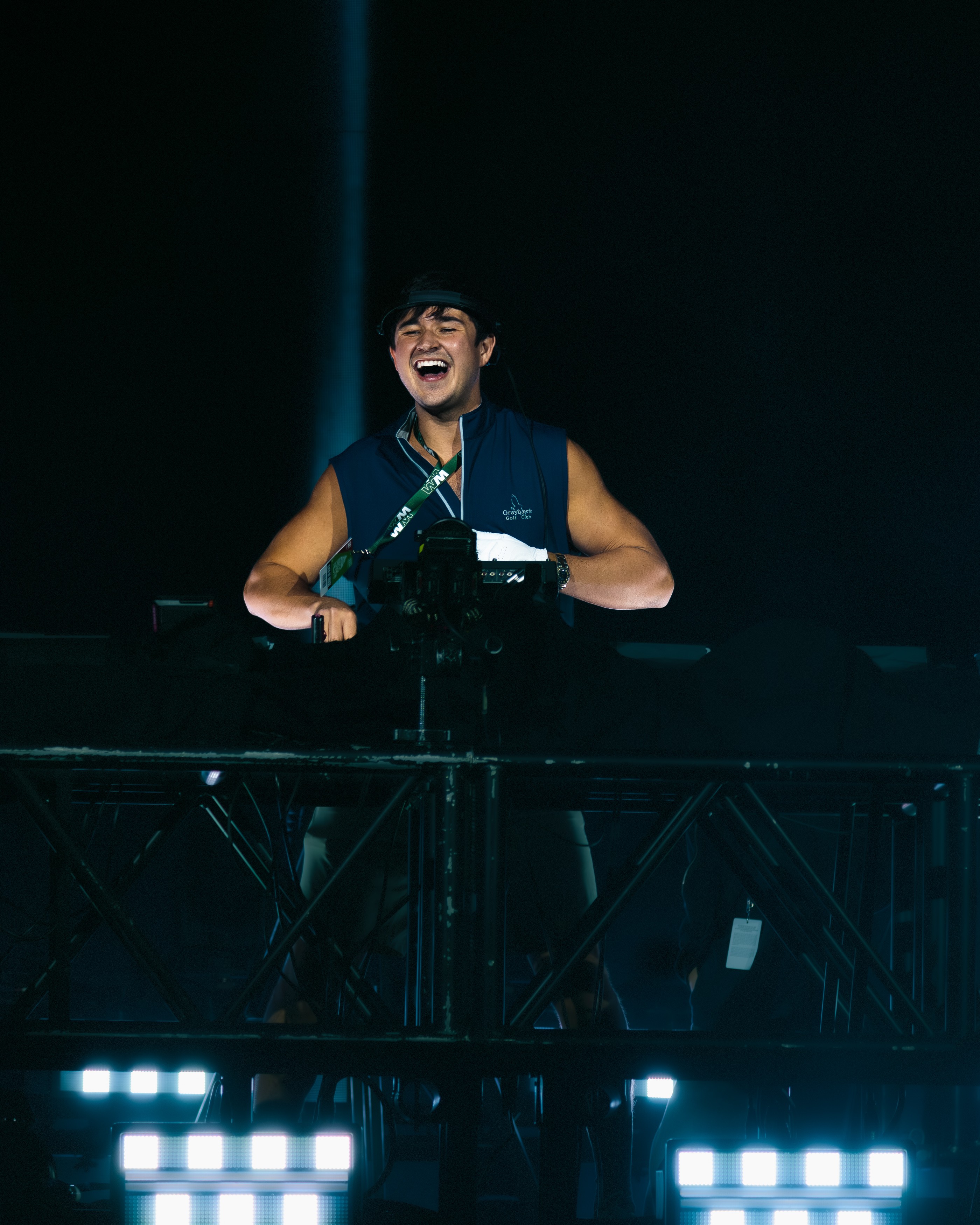 Person smiling and laughing behind a DJ setup with bright lights, dark background, and a spotlight on them.