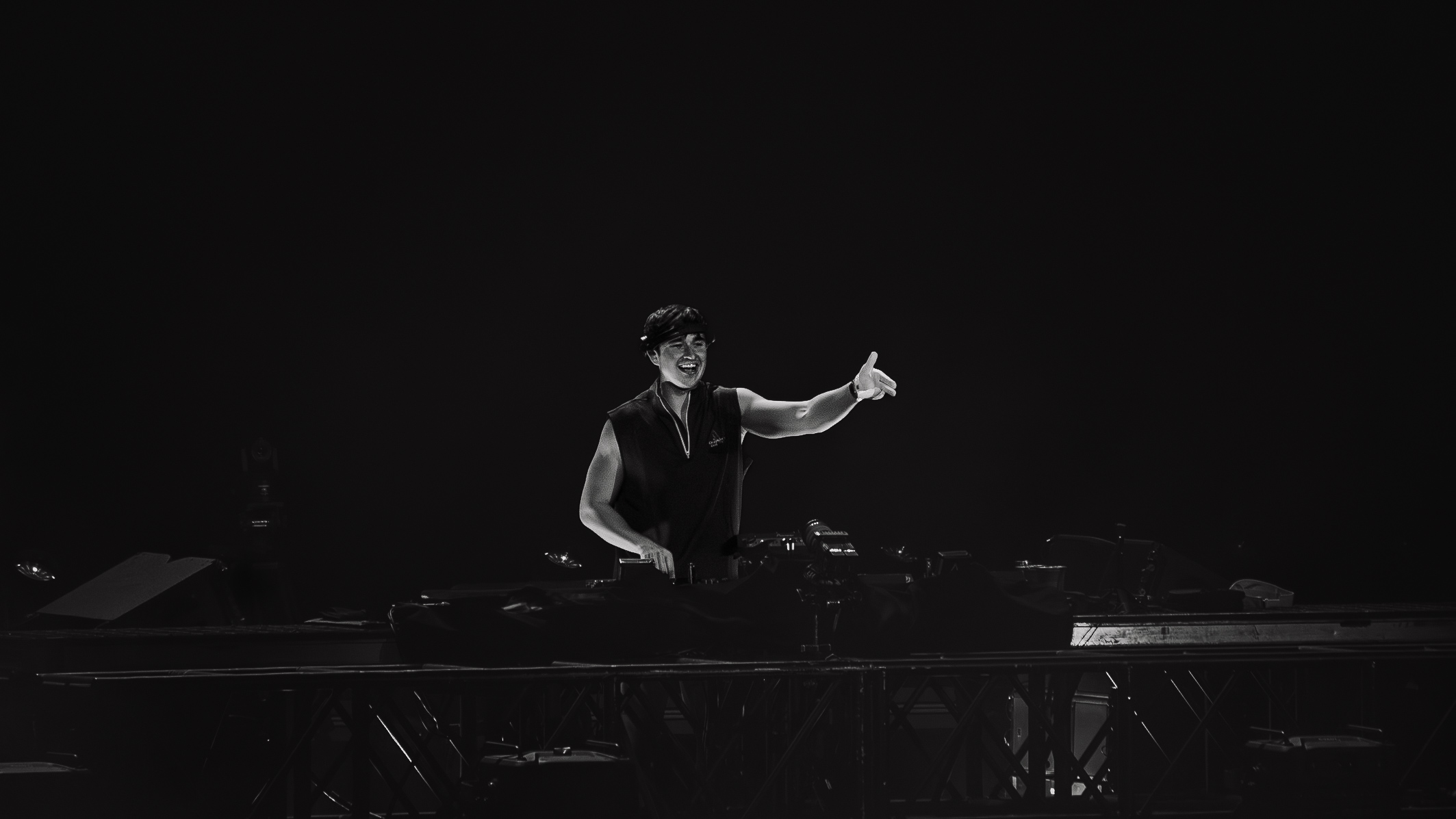 Person standing behind a DJ setup, gesturing with one hand, in a dark environment.