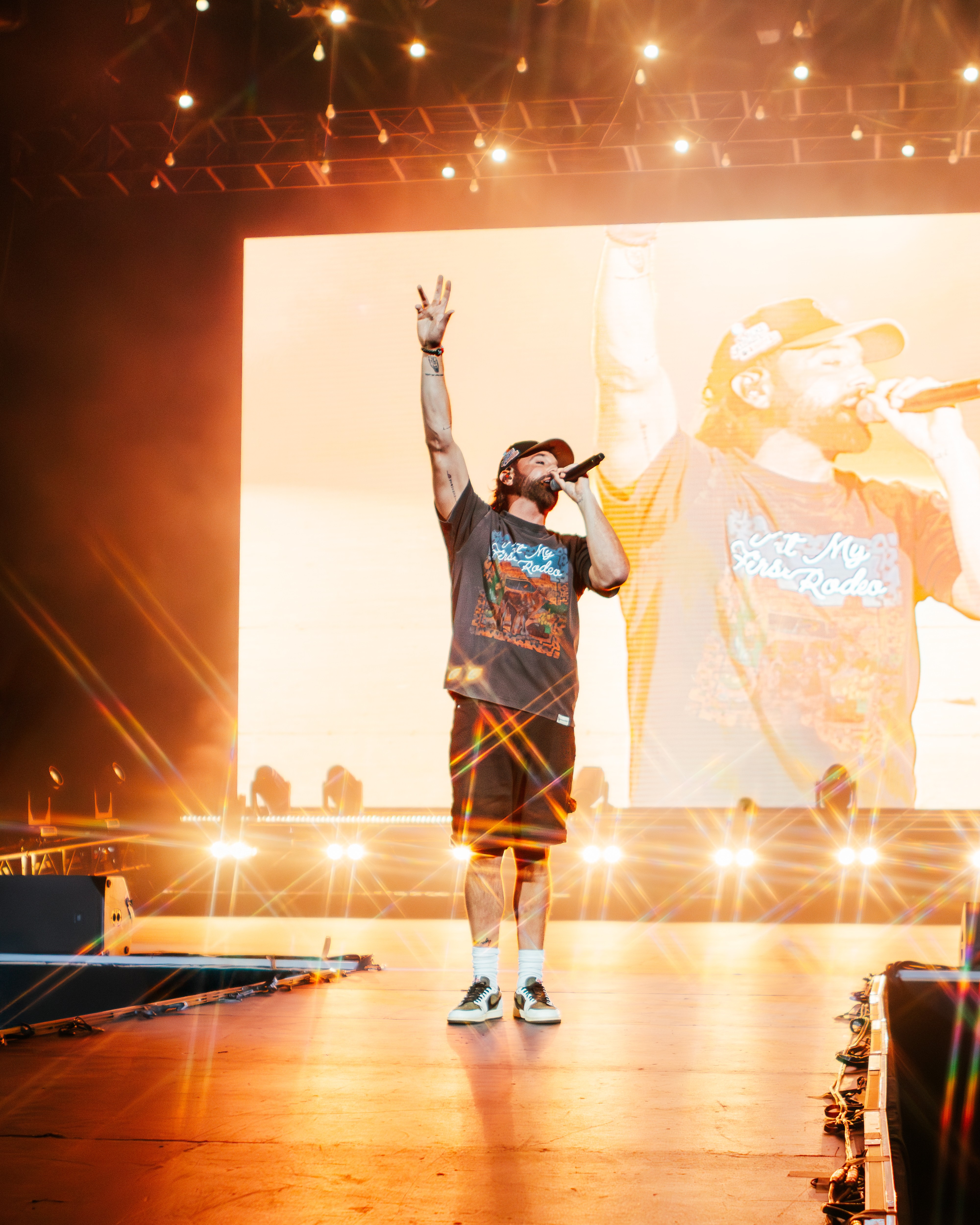 Performer on stage with one arm raised, holding microphone, large screen behind displaying the same image, stage lights shining.