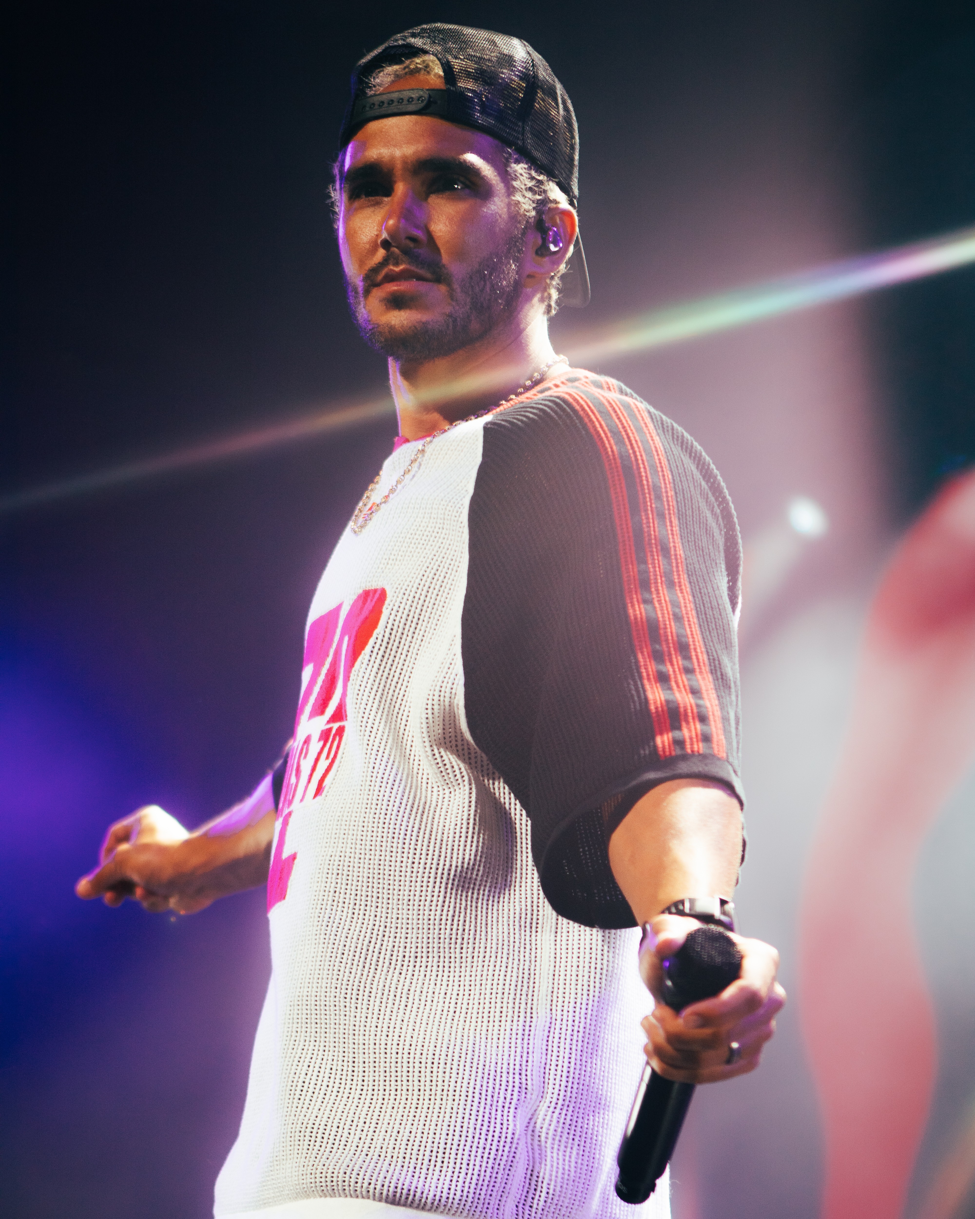 Man wearing a cap and sports jersey holding a microphone on stage with colorful lighting.