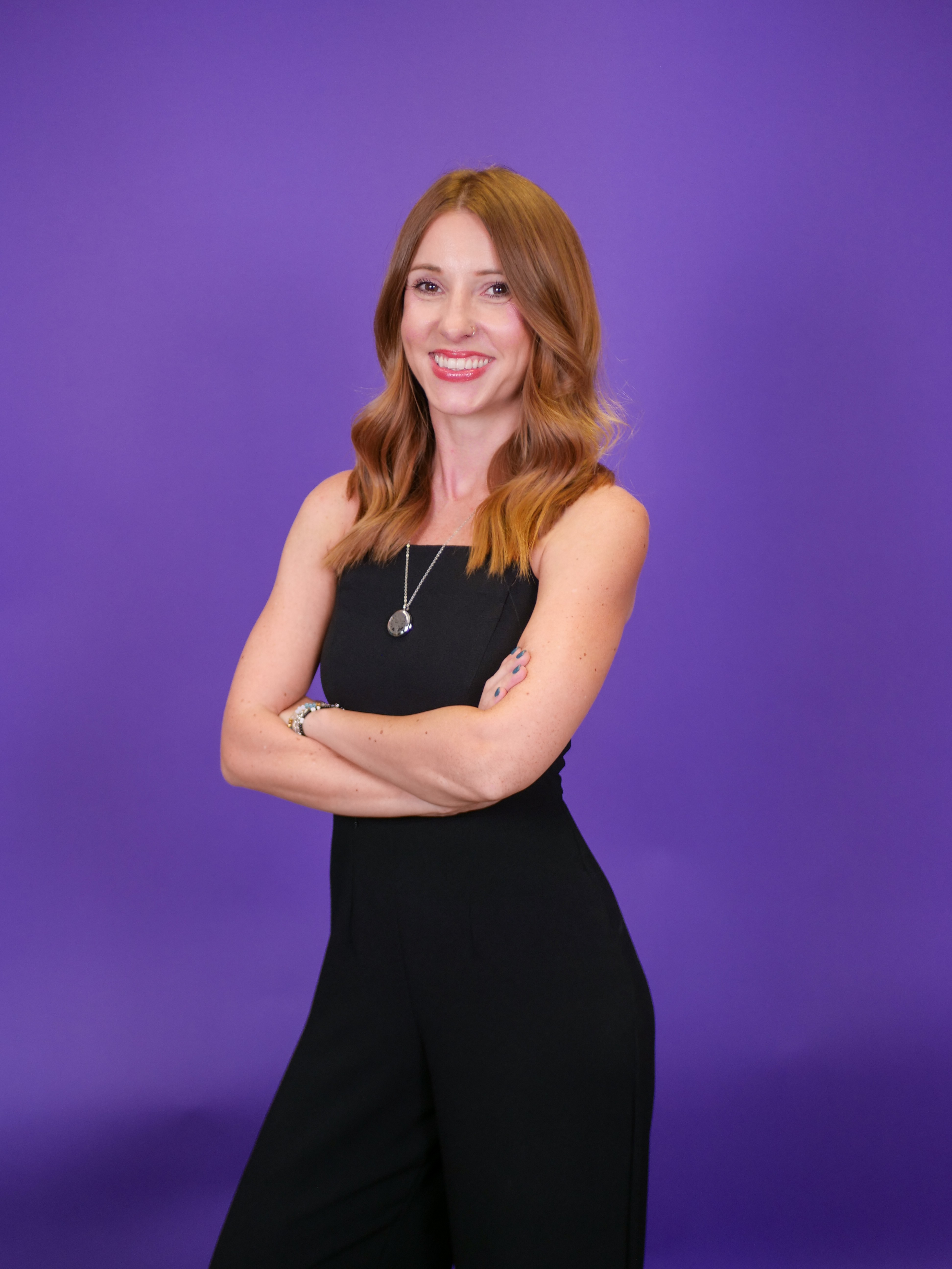 Woman with wavy red hair smiling, arms crossed, wearing a black sleeveless dress, standing against purple background.