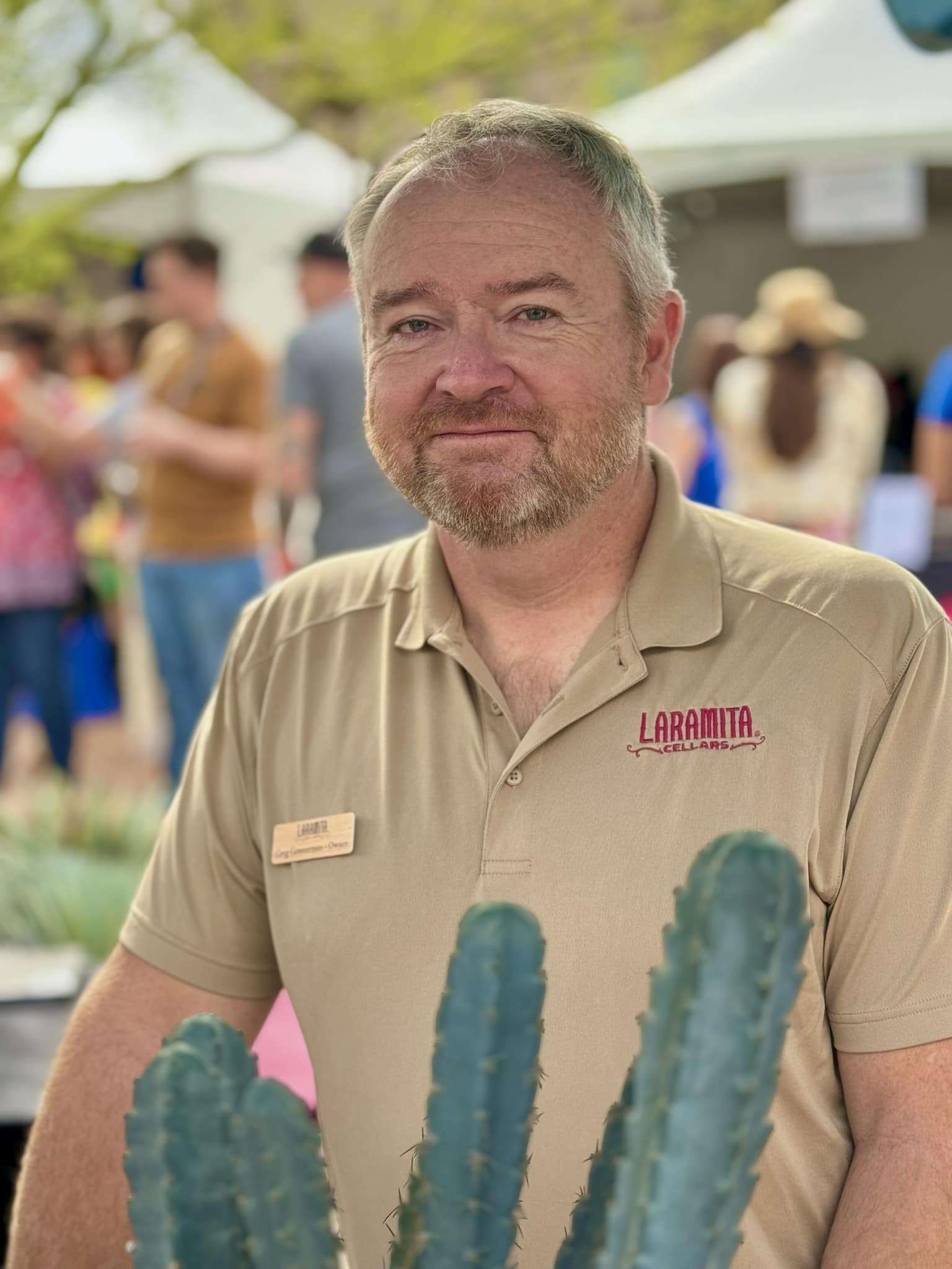 Man with short gray hair and beard wearing a beige shirt with a logo, outdoors at event with people and tents.
