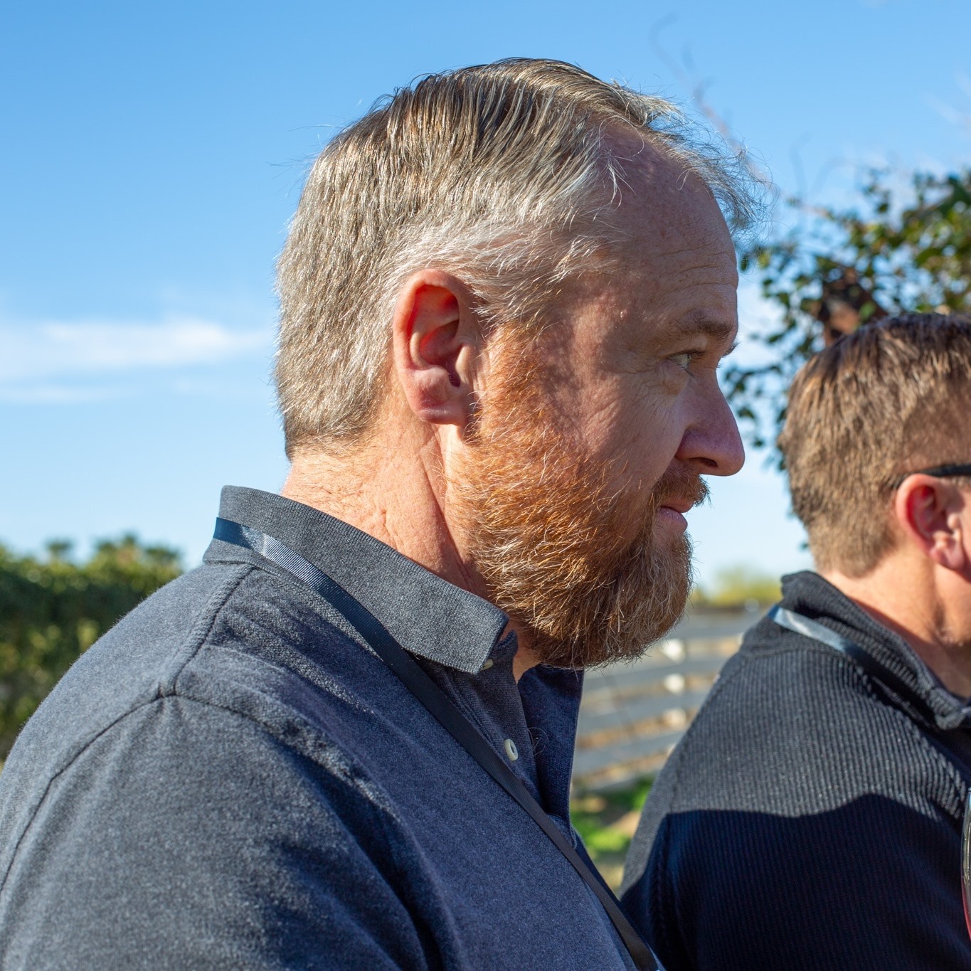 Side profile of a man with a beard and gray hair outdoors, wearing a dark jacket, with a blue sky background.