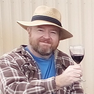 Man wearing a hat and plaid shirt holding a glass of red wine, smiling, in front of a beige background.