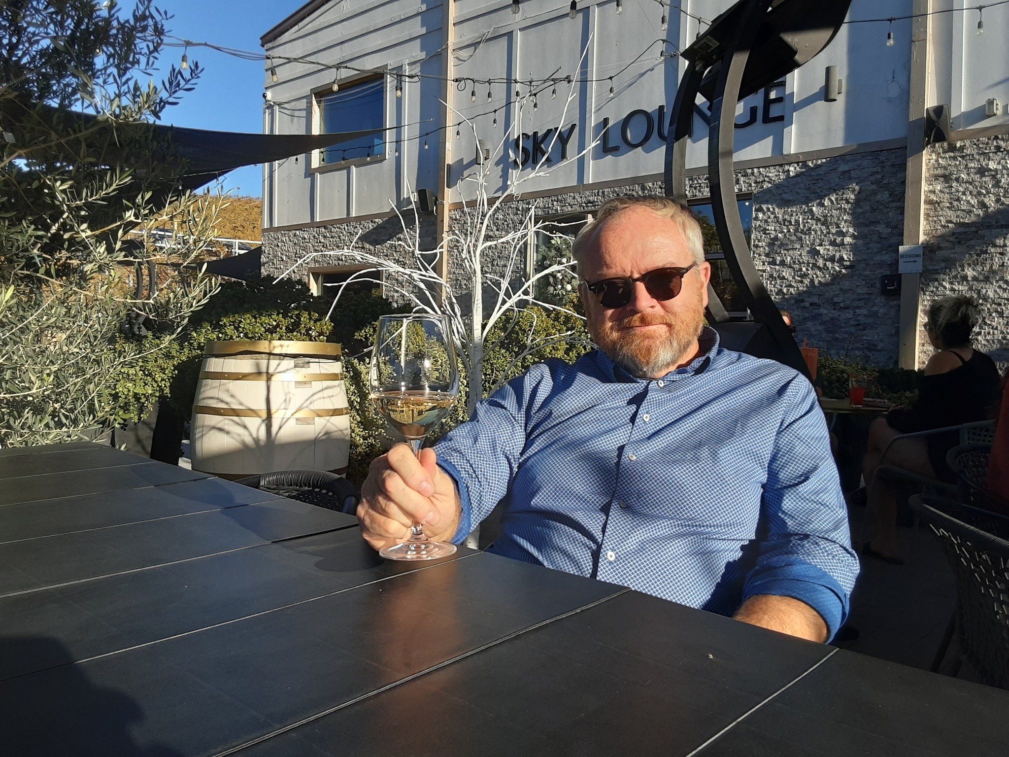 Man wearing sunglasses and a blue shirt sitting at an outdoor table with a drink.