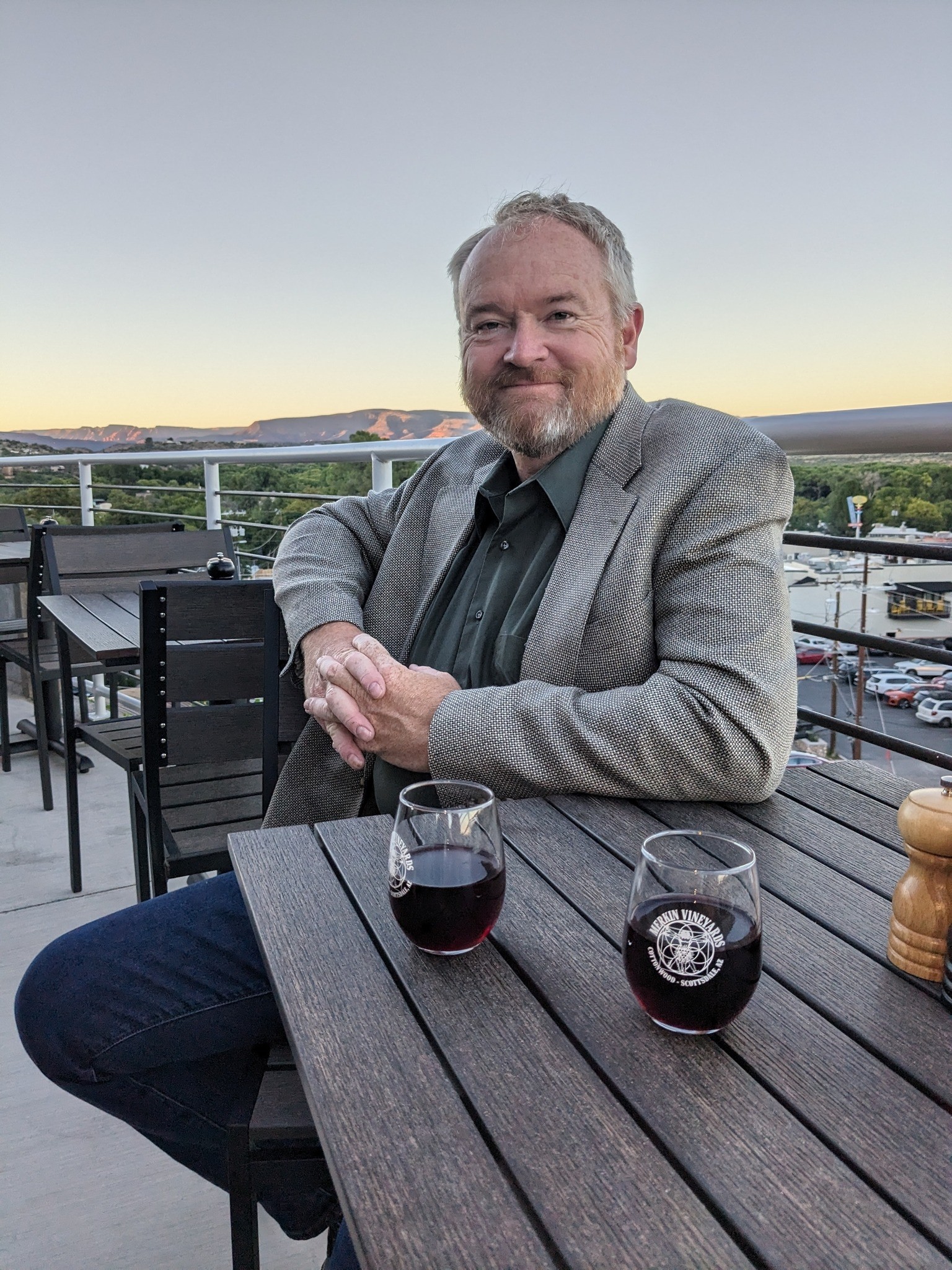 Man with gray hair and beard sitting at a table outdoors with two glasses of red wine, smiling.