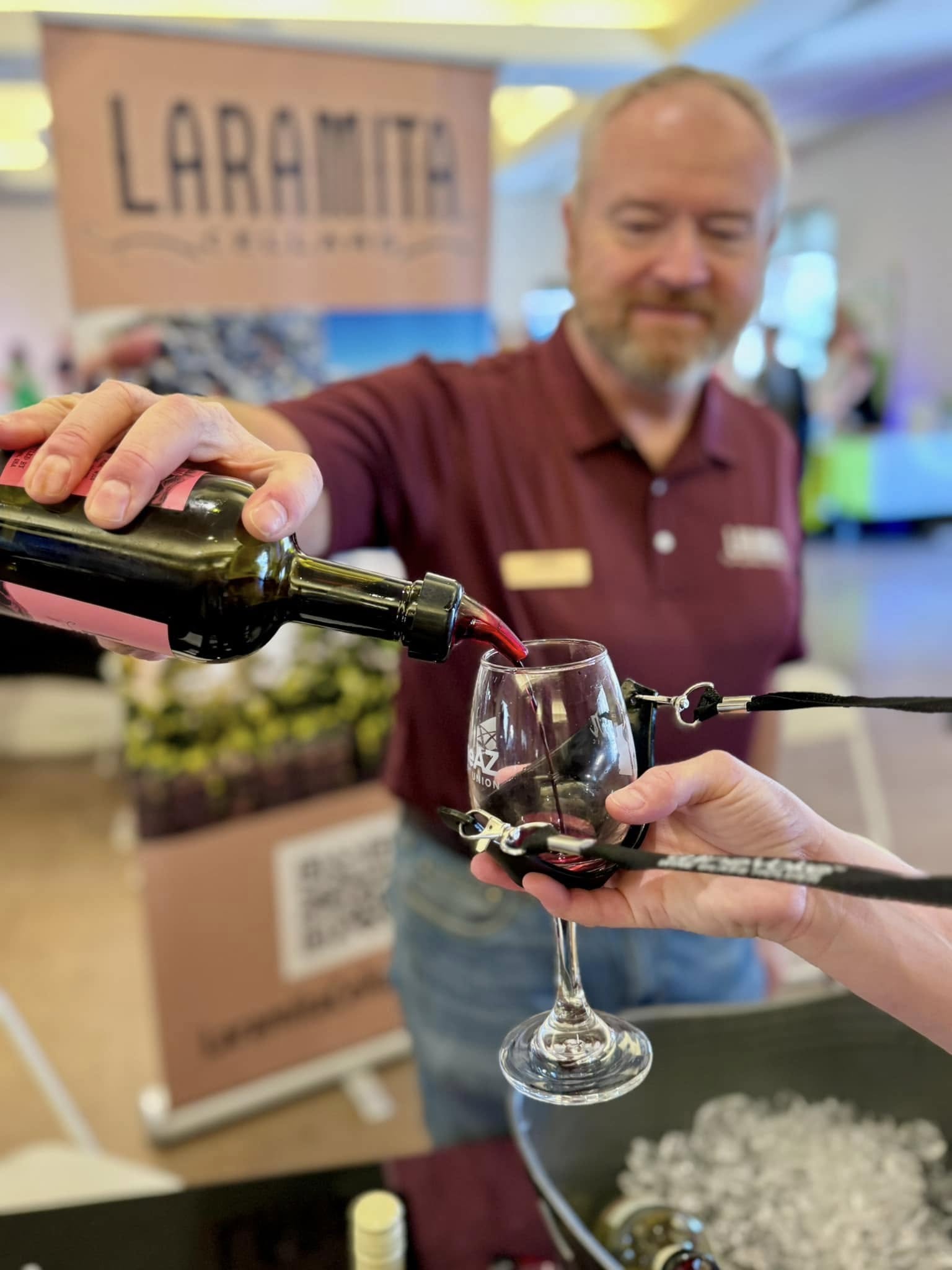 Person pouring red wine into a glass at an event with a Laramie sign in the background.