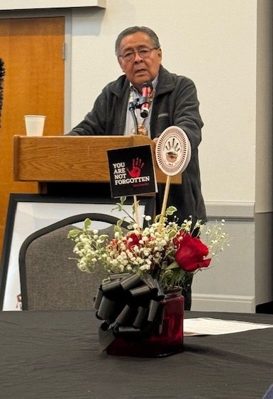 Man speaking at a podium with a microphone, in a room with a table and flower arrangement in the foreground.