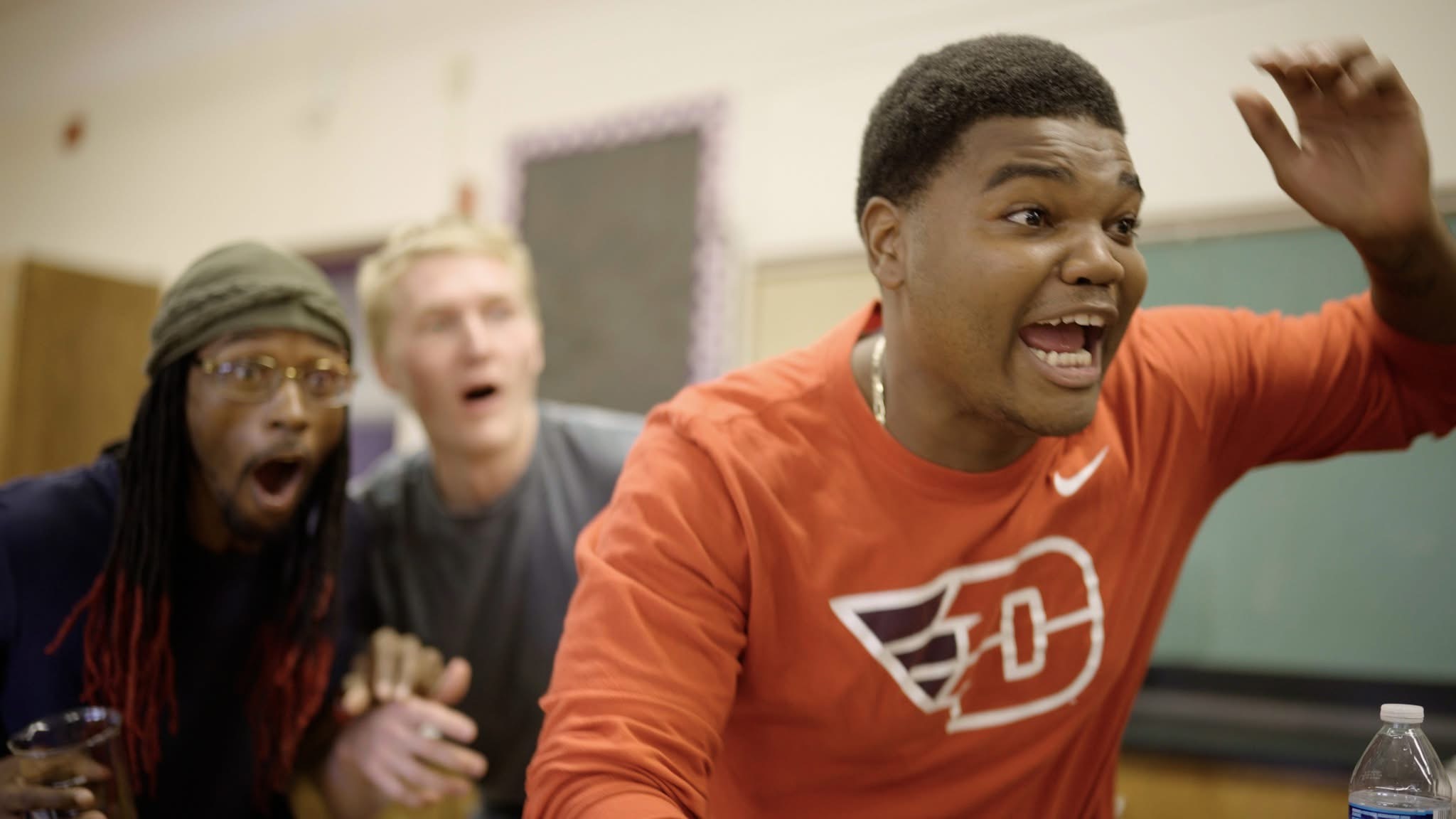 Three people with surprised expressions, one in the foreground raising hand, in a classroom setting.