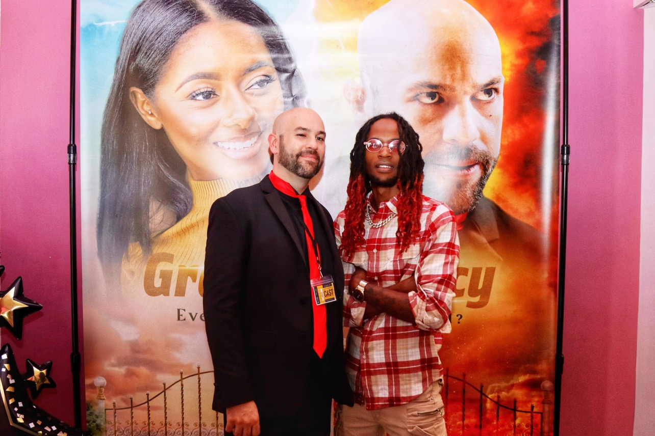 Two men stand in front of a large poster featuring a woman and a man, smiling and posing for the photo.