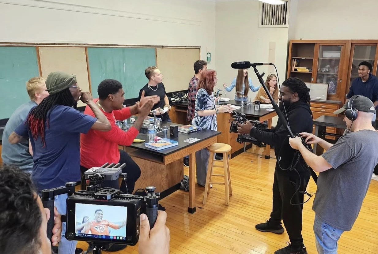 Group of people in a classroom setting, some taking photos or videos, others watching a person with a camera.