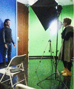 Two people in a studio with green and blue walls, one standing near a chair, the other near a large light, facing each other.