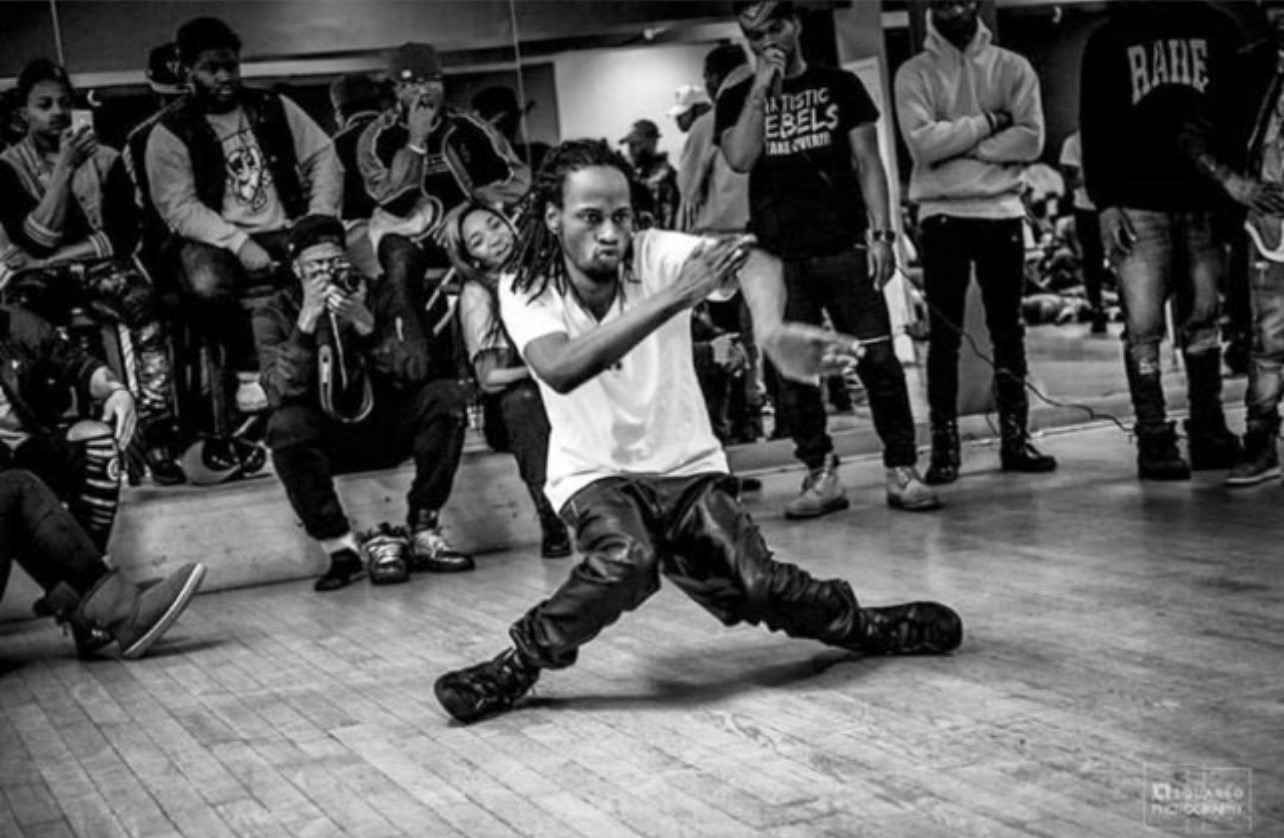 Person performing a dance move on a wooden floor with a group watching in the background.
