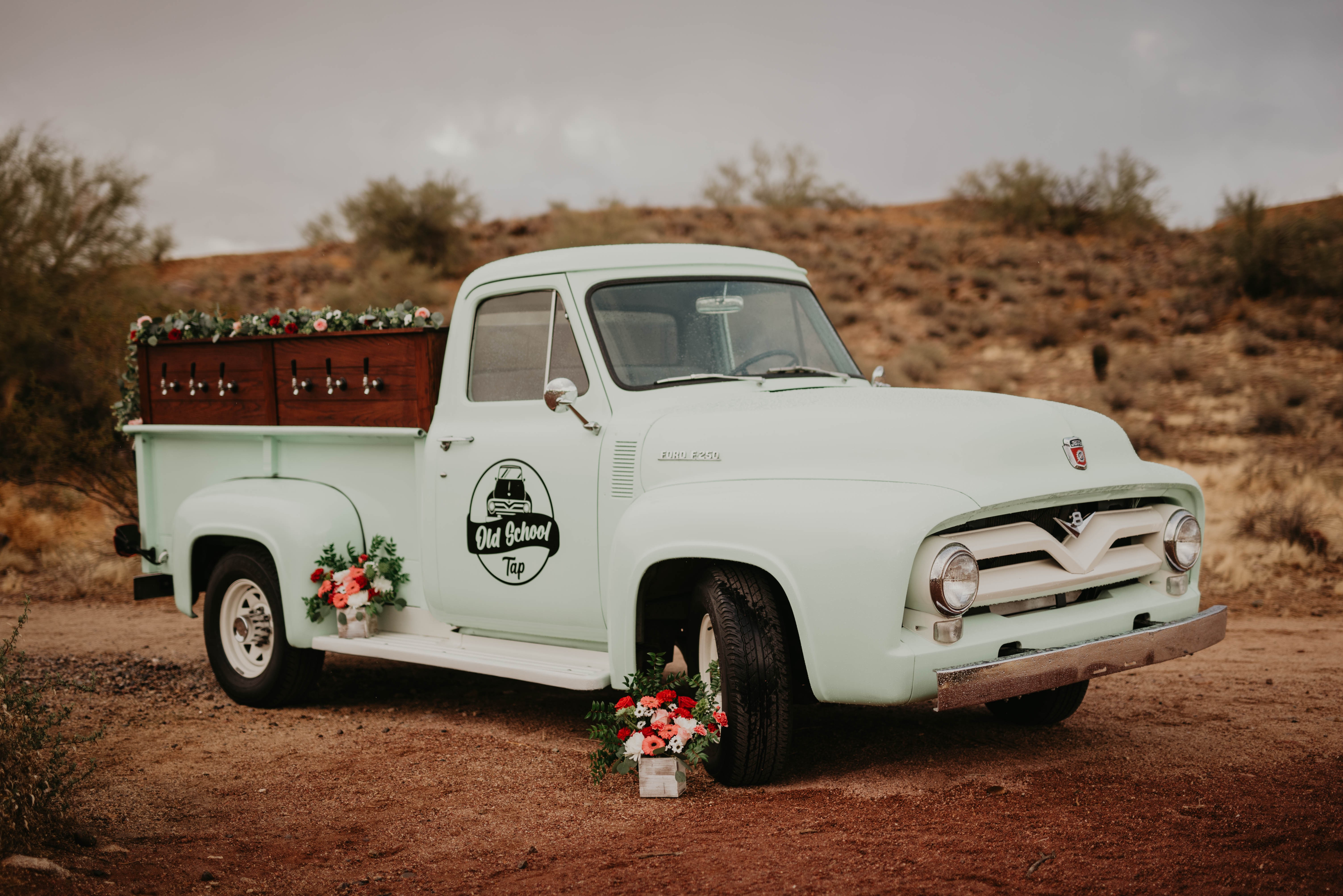 Vintage white pickup truck with floral decorations, parked on dirt with desert landscape in background.