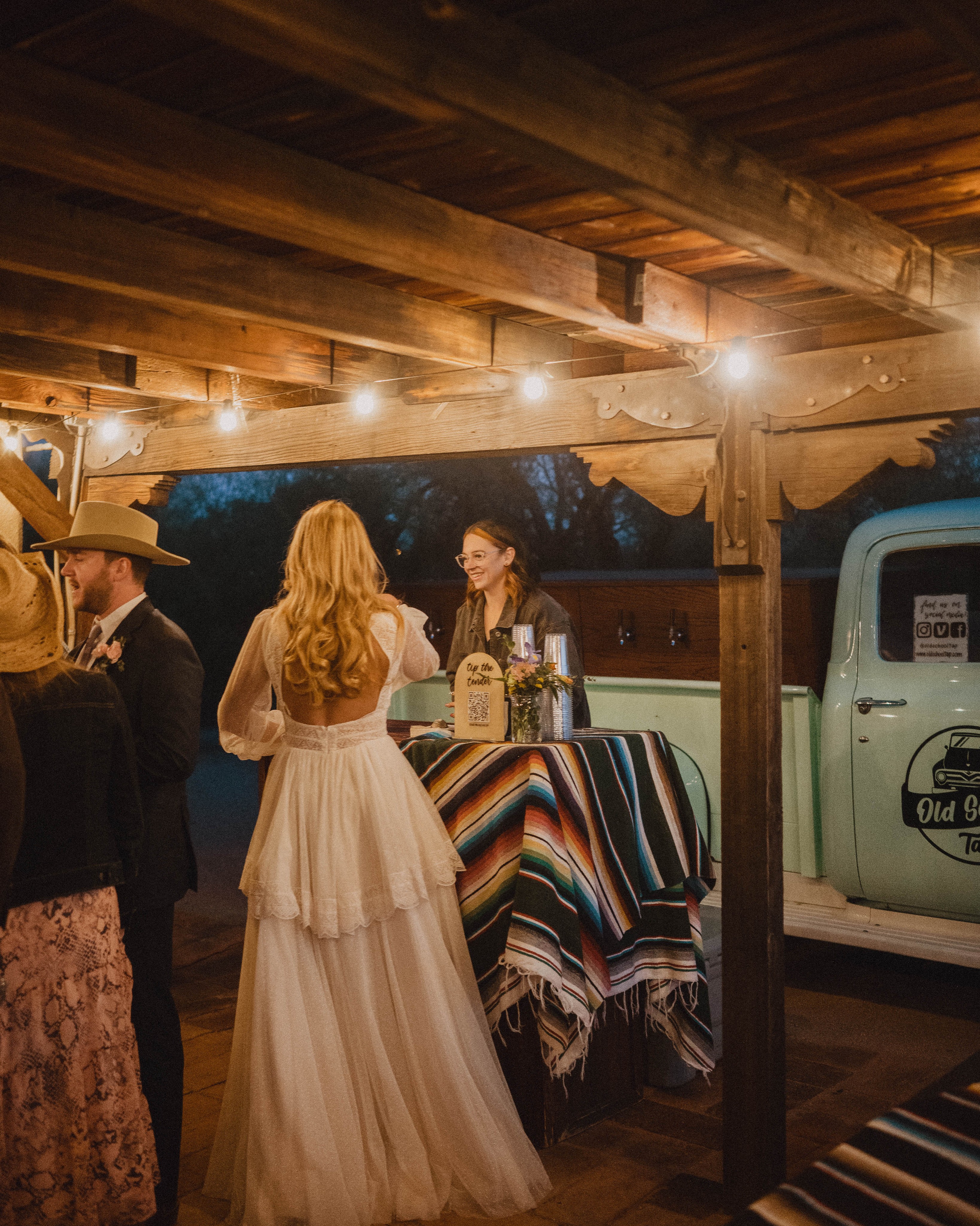 Woman in a long white dress standing at a colorful table with a woman behind it, under a wooden ceiling.