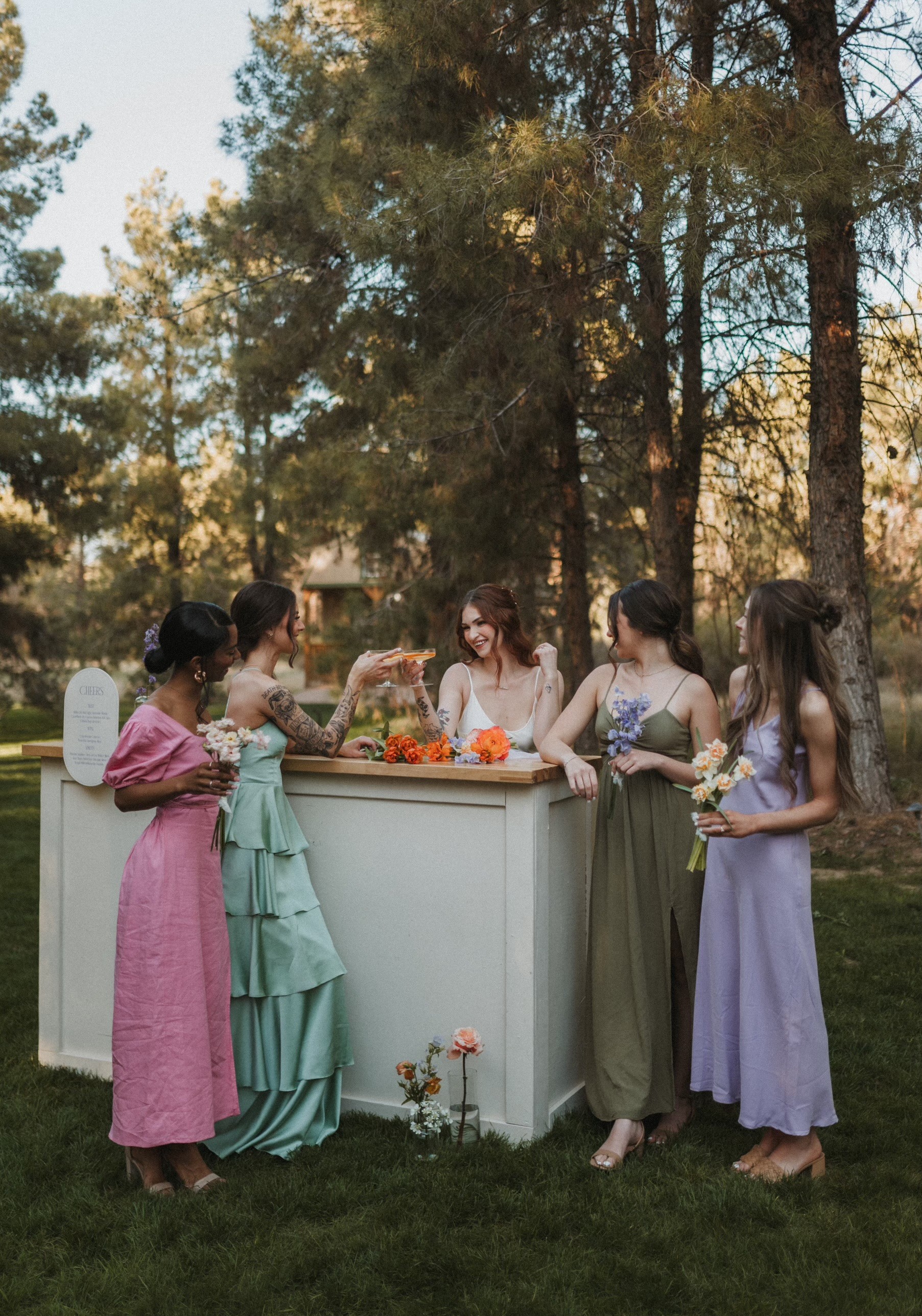 Five women and a child gather around a white bar in a park with trees, flowers, and sunlight.