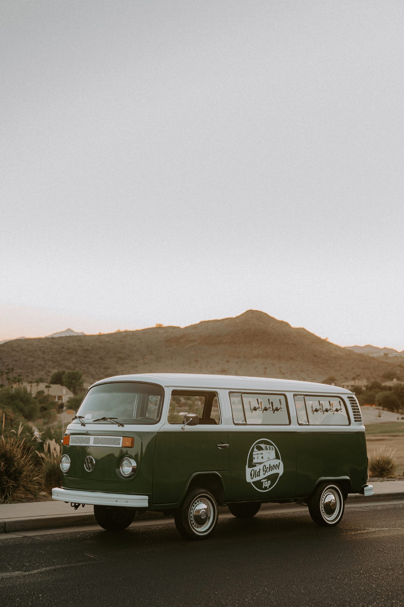 Green vintage van parked on road with desert landscape and hills in background.