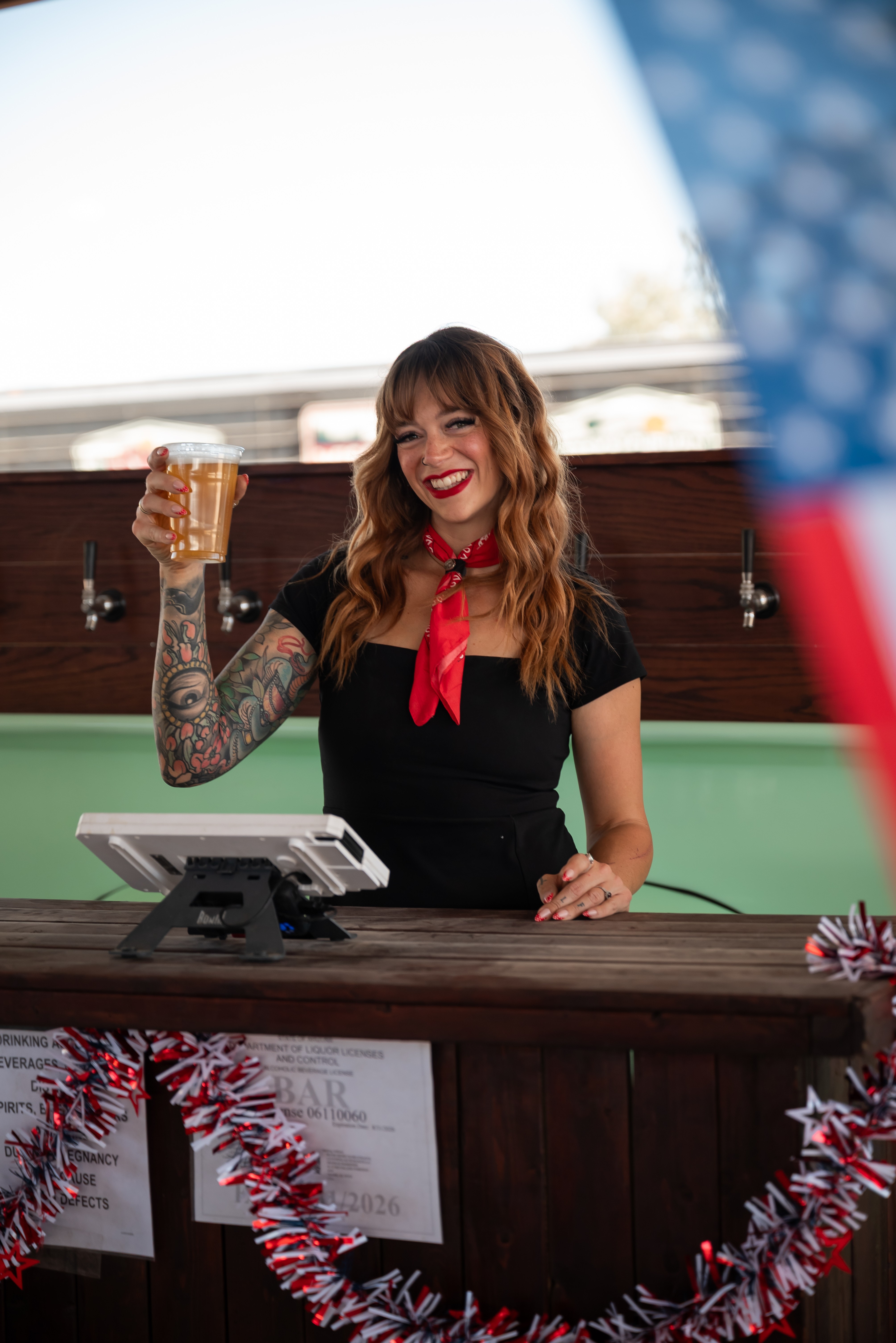 Woman with long hair, tattoos, and red scarf smiling, holding a drink, standing behind a bar counter decorated with garland.