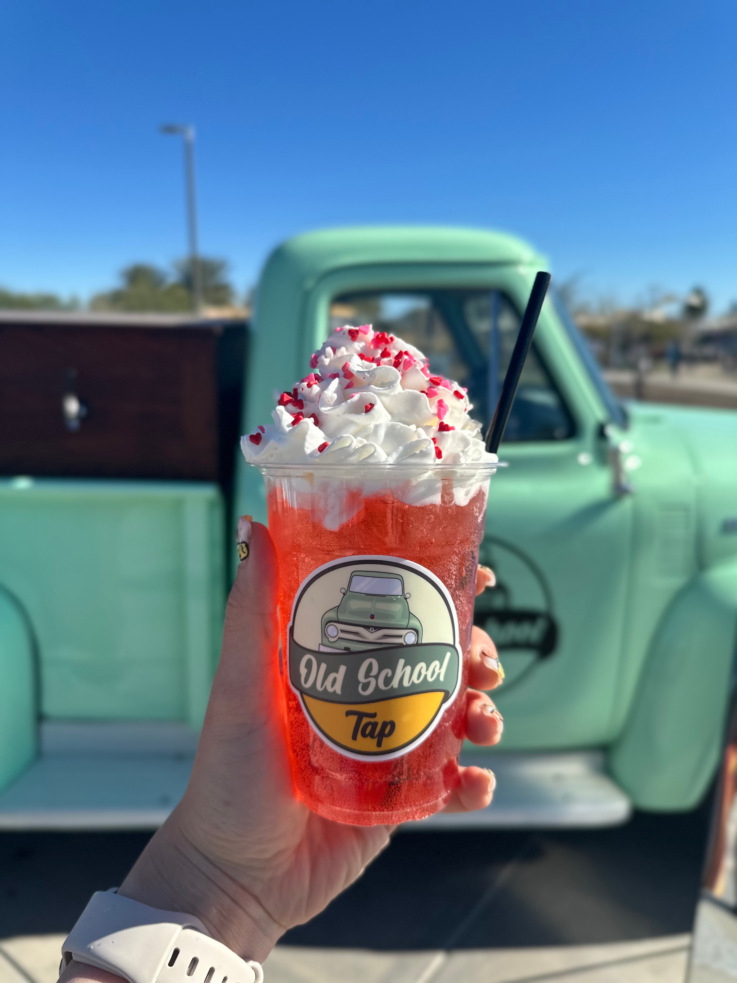 Hand holding a red drink with whipped cream and sprinkles in front of a vintage green truck.