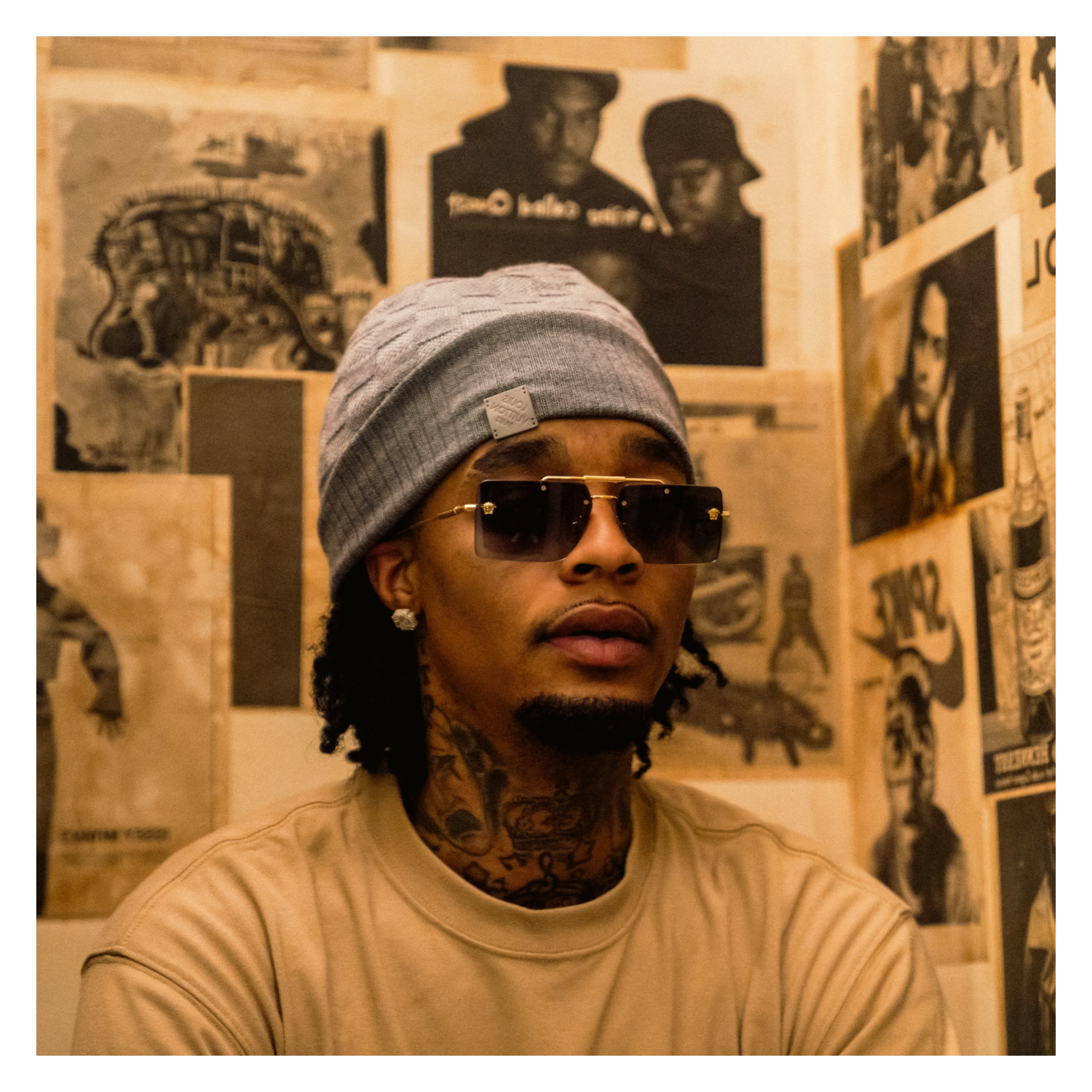 Person wearing sunglasses, gray beanie, and beige shirt in a room with posters on the wall.