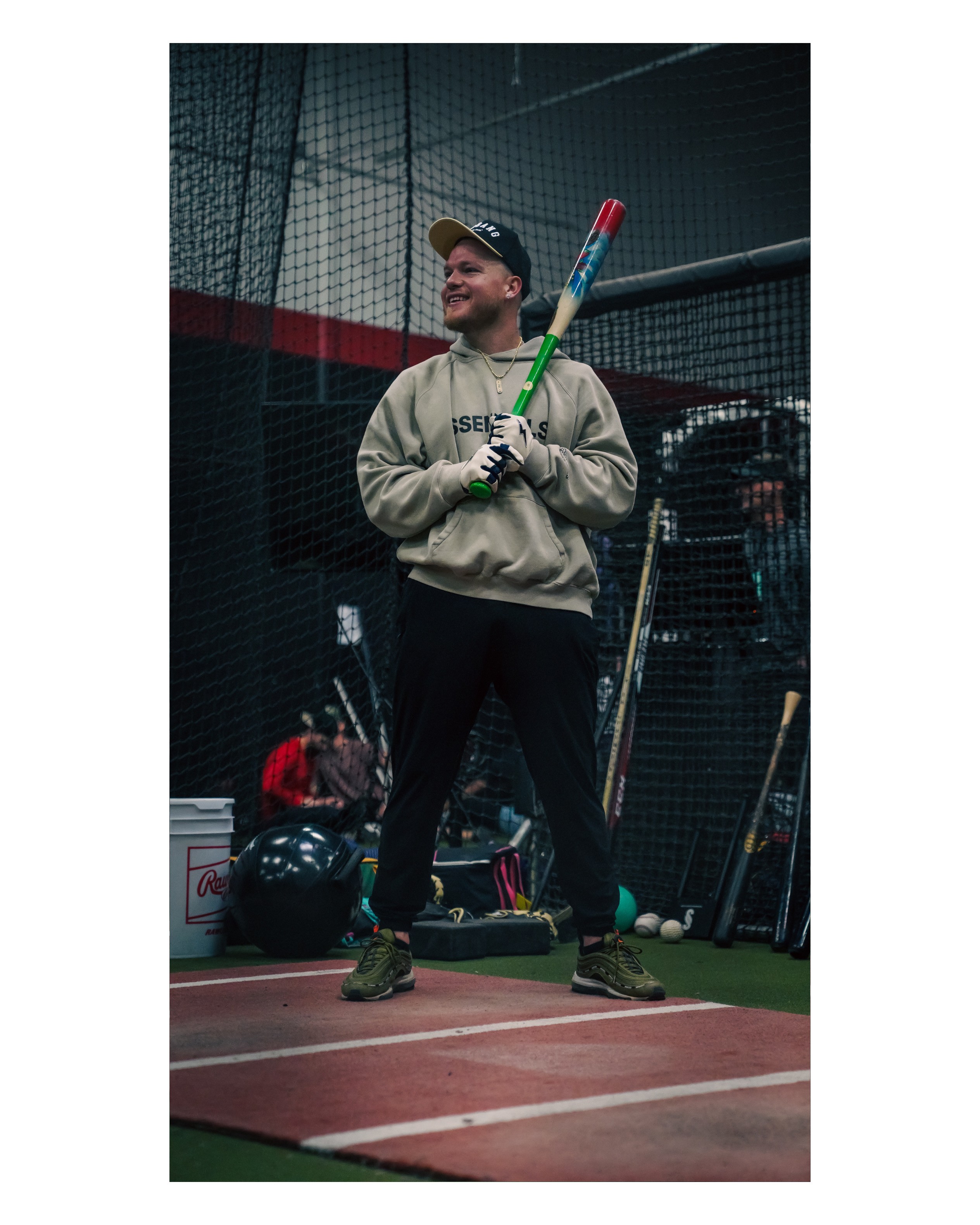 Person standing on indoor baseball field holding a bat, smiling, wearing hoodie, cap, and gloves.