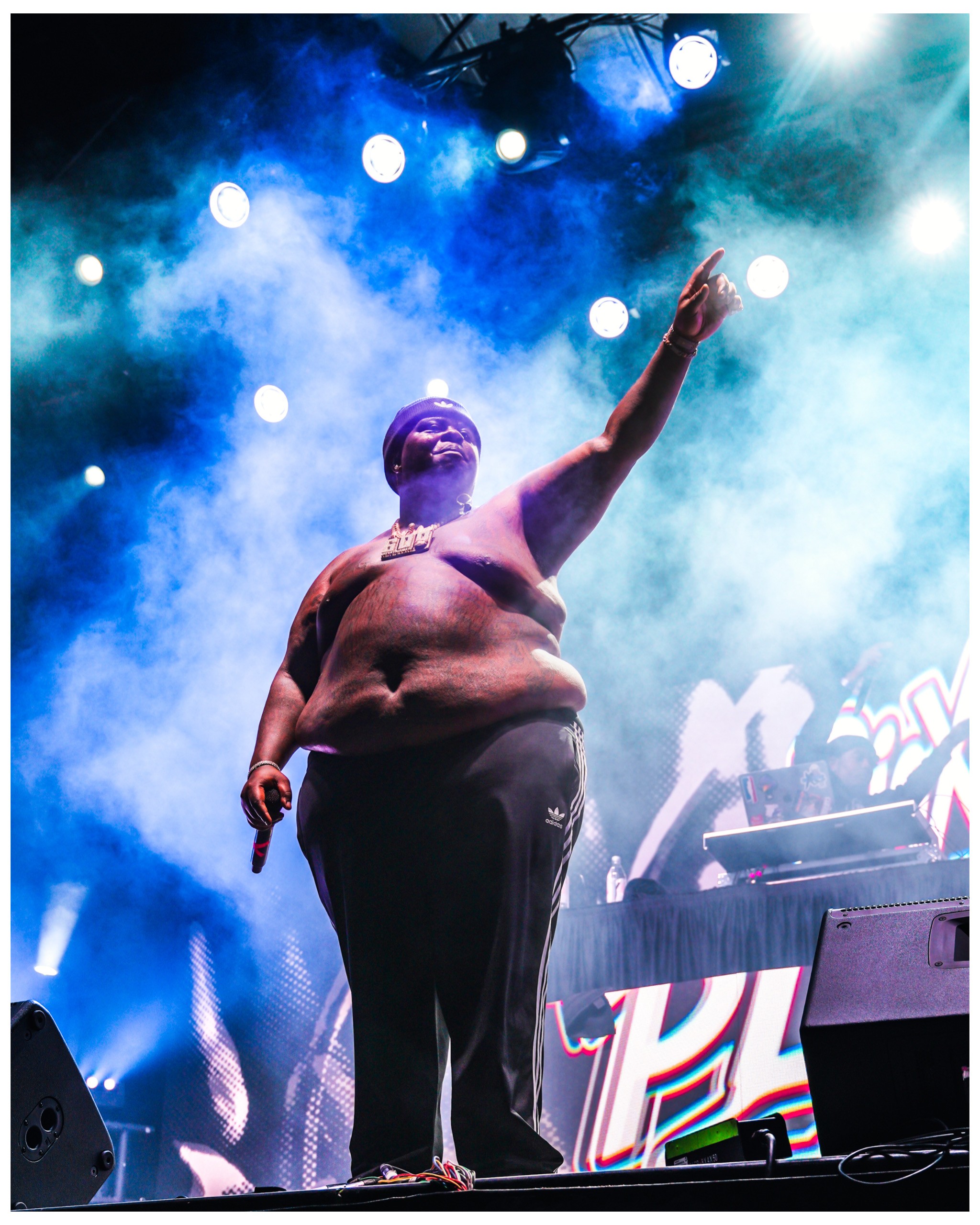 Performer on stage with bright lights and smoke, pointing upward, with a DJ setup in the background.