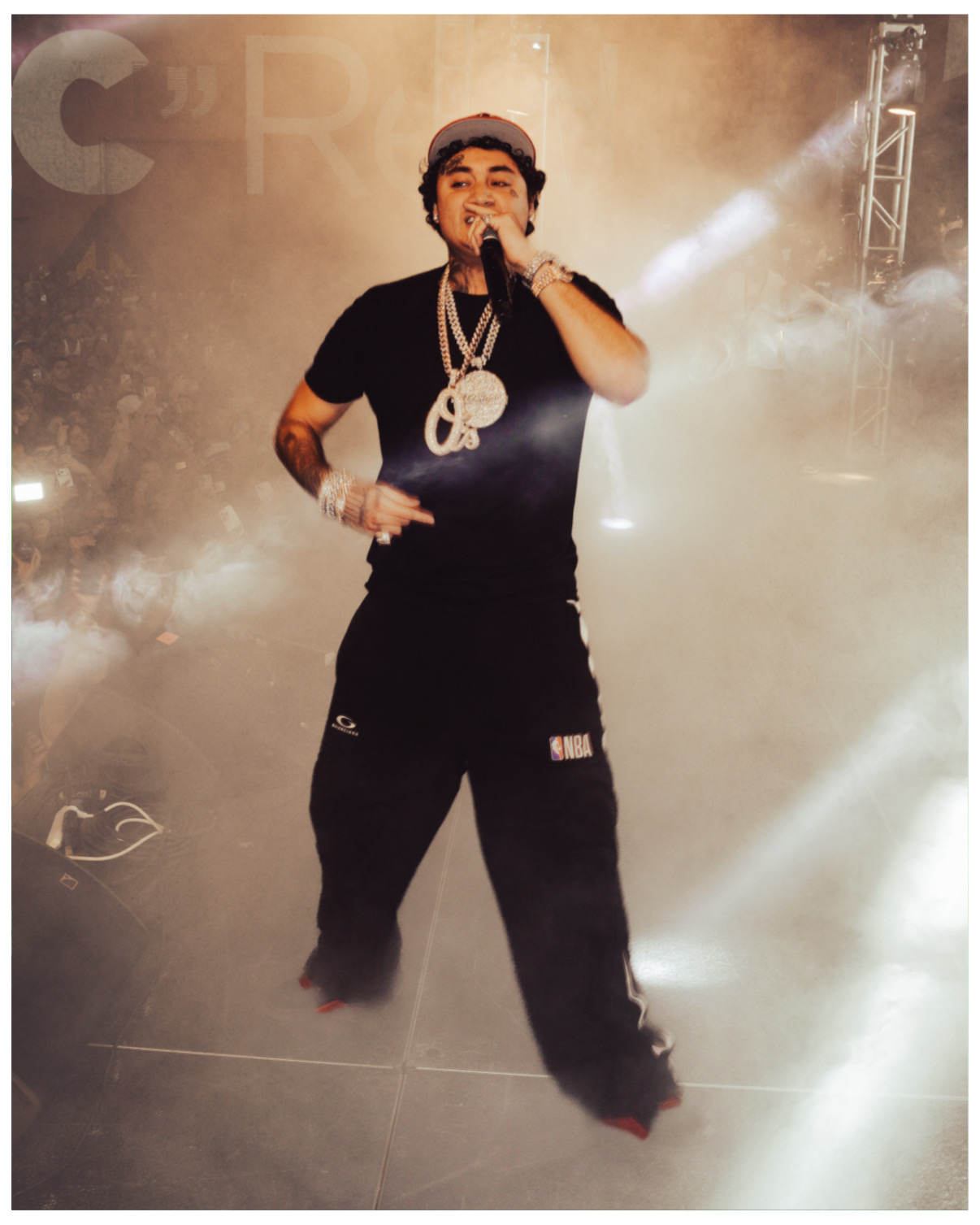 Performer on stage with smoke and bright lights, wearing black clothing and jewelry, holding a microphone.